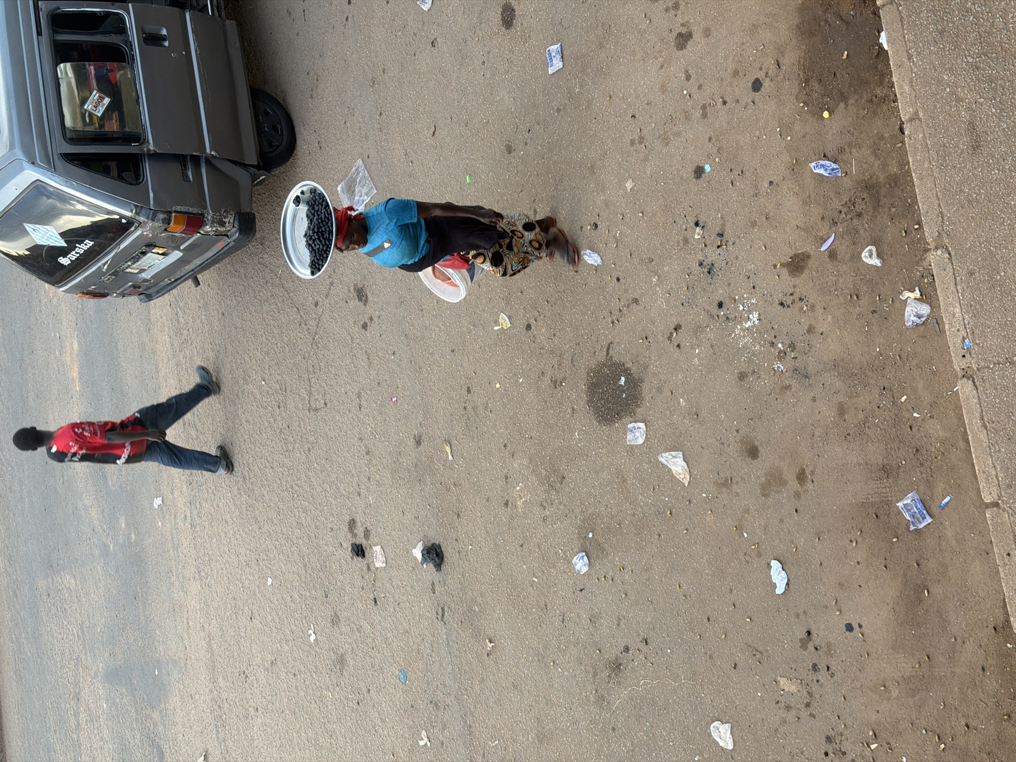 In Bukuru, Nigeria, on a paved road littered with debris, a person walks carrying a tray of dark berries on their head and a bucket in their arm. The person wears a blue top, a red headscarf, and a patterned skirt. To the right, a gray van with open rear doors is parked. Further to the left, another person in a red and black shirt and blue jeans walks away from the van. The ground is predominantly gray asphalt, with scattered litter, including plastic bags and wrappers, and small stones. The lighting suggests it is daytime, possibly late afternoon due to the shadows.