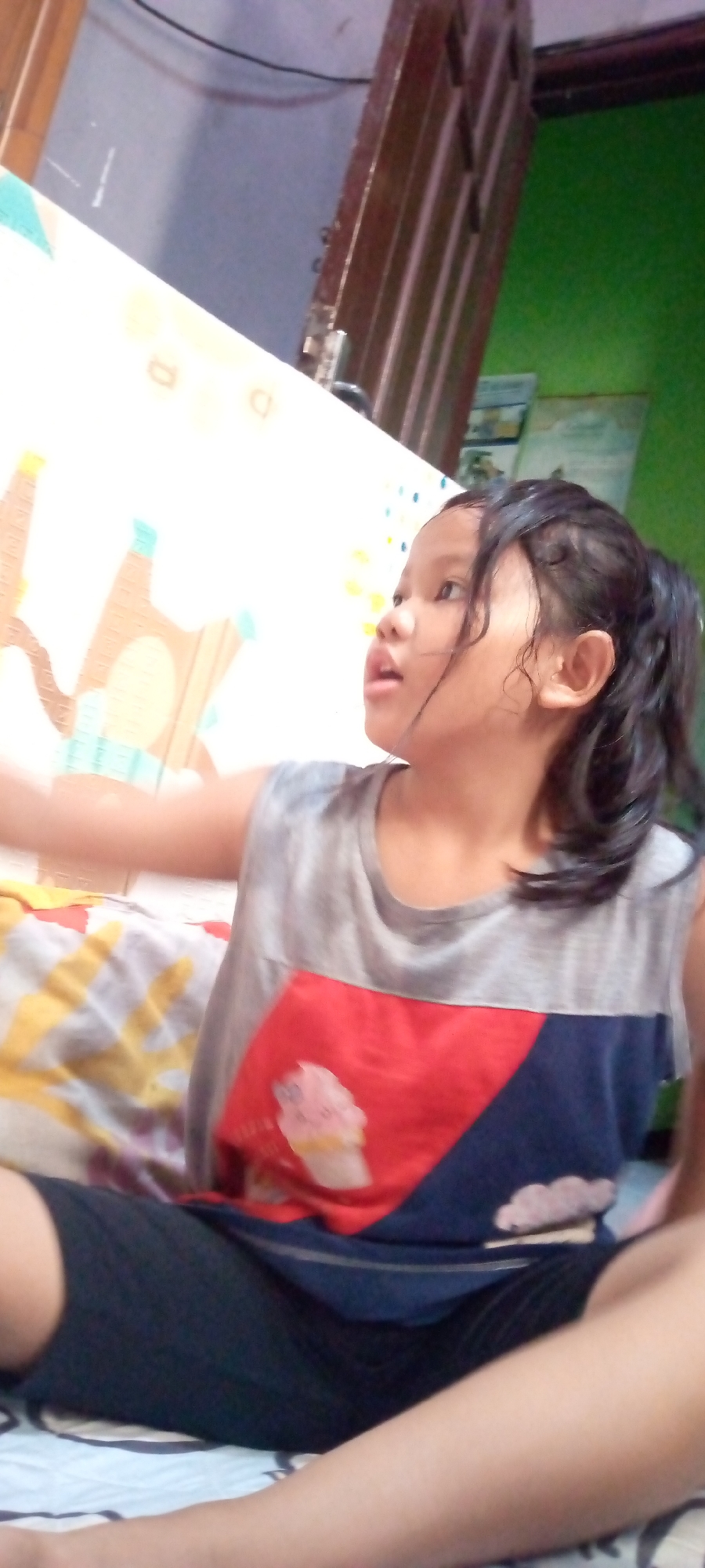 In this indoor image, a young girl is captured from the chest up, sitting on a surface that appears to be a mat or bed. She is wearing a sleeveless shirt with a gray upper portion and a red and navy blue lower portion. The red part of the shirt features a graphic of an ice cream cone. Her dark hair is pulled back into a ponytail and appears slightly damp. The girl is looking to her right with her mouth slightly open, as if she is about to speak or react to something.
The background of the image is blurred but suggests an indoor setting. To the left, a white mat with some colorful patterns is visible. To the right, a wooden structure, possibly a door or frame, and a green wall are discernible. The lighting in the image suggests it was taken during the daytime, likely indoors. The overall impression is a candid shot of a child engaged in an activity or conversation, possibly at home in Sumberwono, Indonesia. No visible text is present in the image.