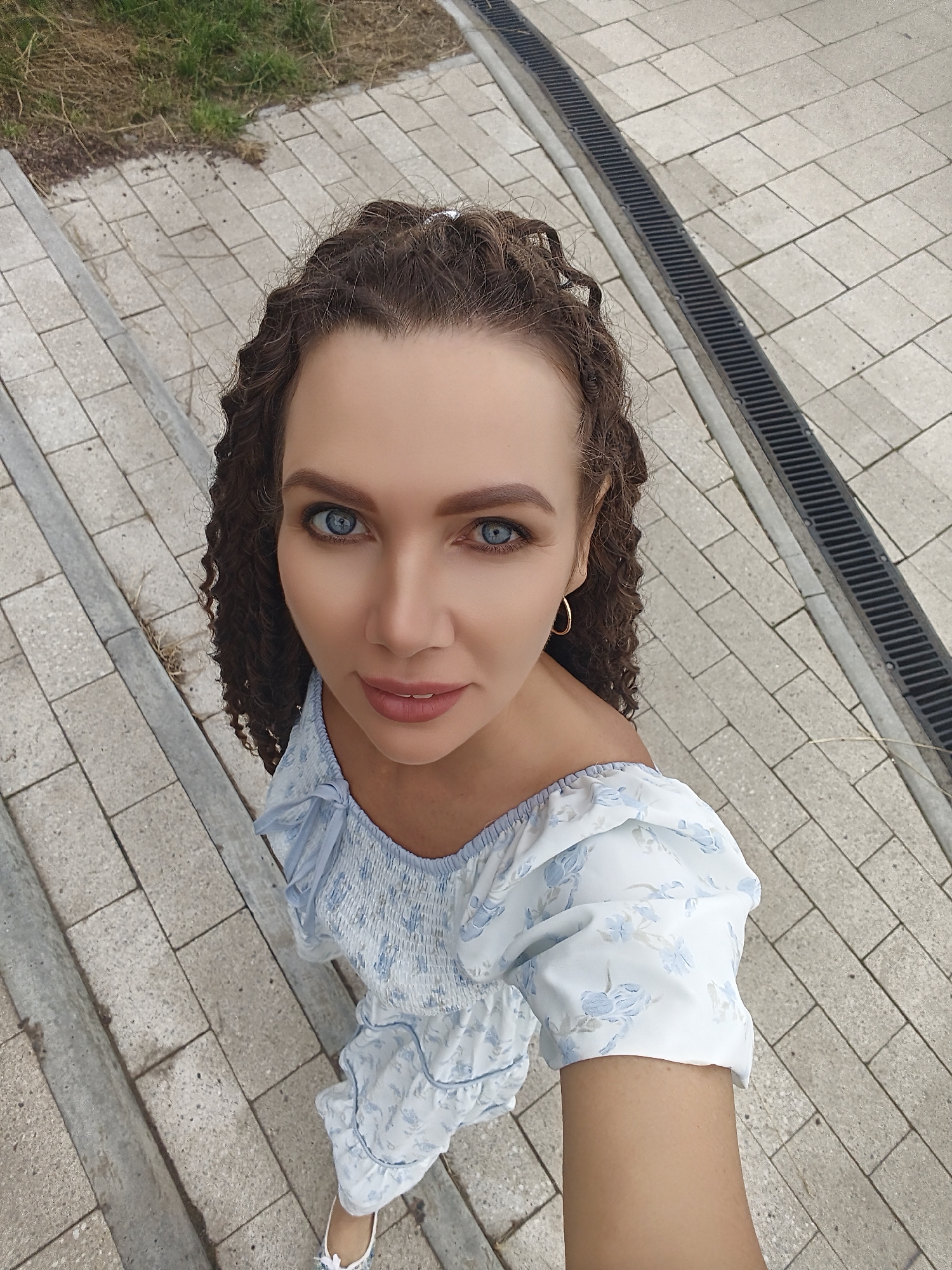 The media file is a medium shot selfie of a woman with curly brown hair and bright blue eyes. She is wearing a white off-the-shoulder dress with a blue floral pattern. The woman is looking directly at the camera with a slight smile. Her left arm is extended, holding the camera.
The setting appears to be outdoors, with a tiled pathway and some grass visible in the background. There is also a drainage grate running along the side of the path. The lighting suggests it is daytime, and the weather appears to be overcast.
The woman's expression is neutral to pleasant, conveying a sense of calm or contentment. No text is visible in the media file. The location is identified as Chaikovsky, Russia.