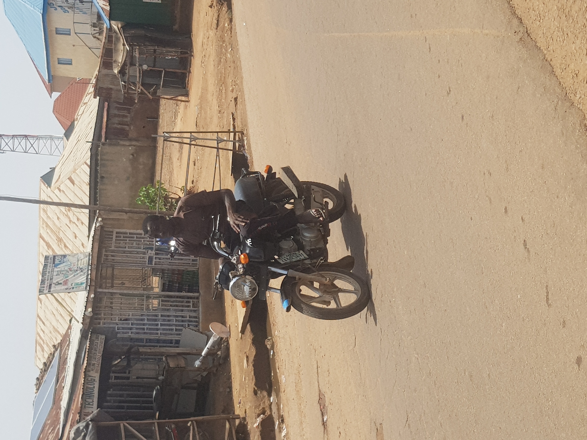 In Tukur Tukur, Nigeria, a Black man is riding a blue and silver motorcycle on a paved road. He is wearing a dark-colored shirt and dark pants. The motorcycle has a license plate that is partially visible. In the background, there are several buildings with corrugated metal roofs, some of which appear to be shops or small businesses. One building has solar panels on its roof. A tall telecommunications tower is visible in the distance. The environment appears to be an urban or semi-urban area with a dry, dusty atmosphere.