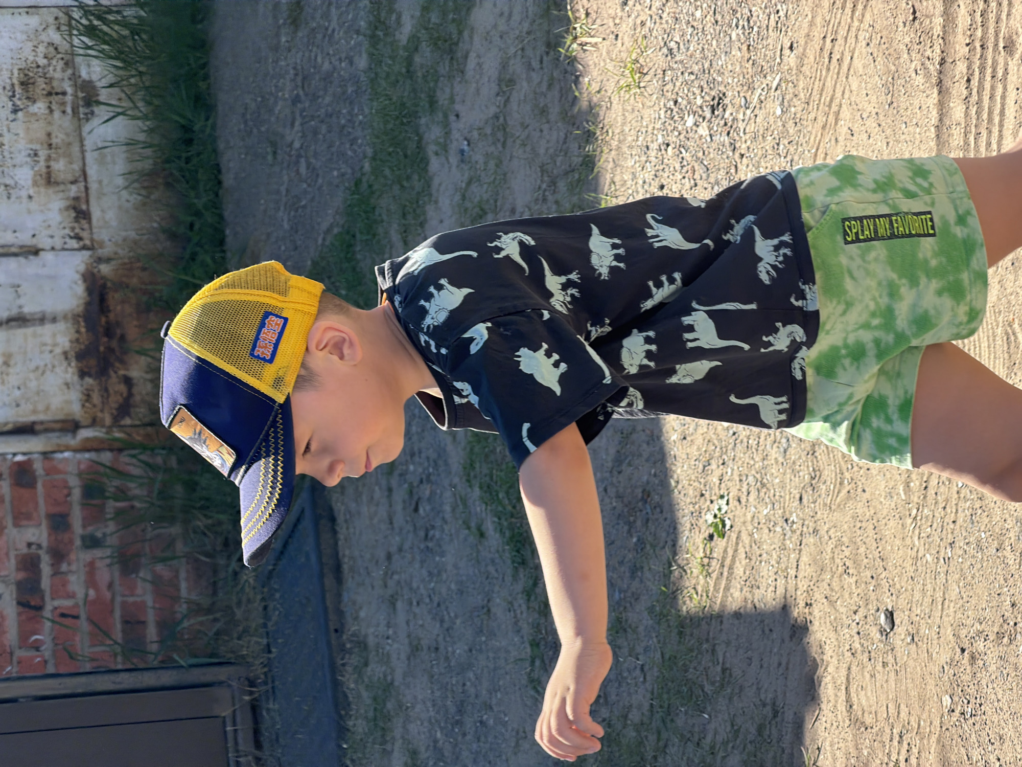 A young child is seen in an outdoor environment. The child is wearing a black shirt with white dinosaur prints, green and white tie-dye shorts with "SPLAY MY FAVORITE" printed on them, and a two-toned blue and yellow cap. The child appears to be walking or reaching down with their right arm extended. The ground is gravelly, with some grass and vegetation near the edges. In the background, there is a brick structure or wall. The photo appears to be taken in the city of Chaikovsky, Russia.