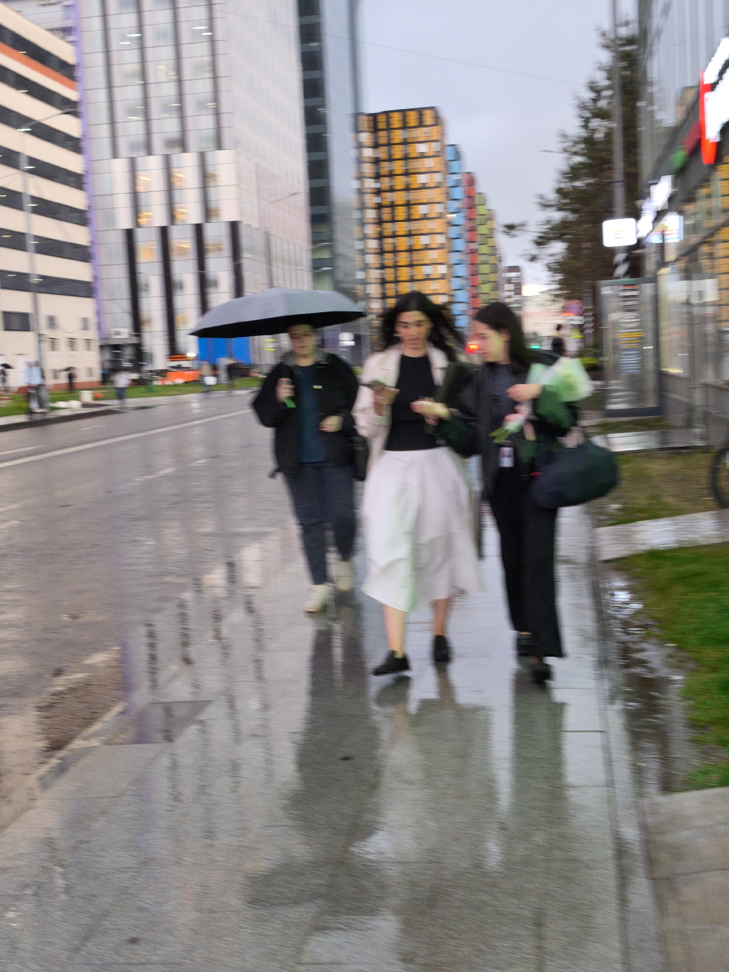 The photo depicts three individuals walking on a sidewalk, likely in Moscow, Russia, evidenced by the presence of Moscow City skyscrapers in the background. It appears to be a rainy day, with wet sidewalks reflecting the surroundings. The first individual is holding a black umbrella. The second individual is wearing a white skirt and appears to be holding a phone. The third individual is carrying what appears to be a bouquet of flowers. The general impression is one of a city street scene on a cloudy day.