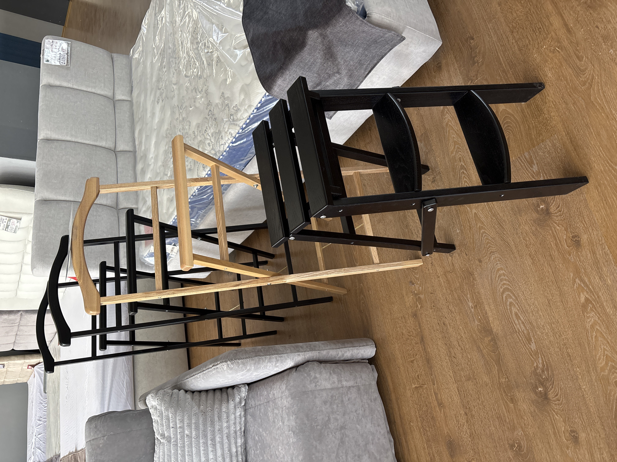 The image depicts a retail environment, likely a furniture store, with a focus on beds and seating. In the foreground, there are three folding step stools arranged in a diagonal line. The stool closest to the viewer is black and has three steps. Behind it is a light-colored wooden step stool with two visible steps, and further back, another black step stool is partially visible. The stools are positioned on a wooden floor. In the mid-ground and background, several beds are displayed, some covered in protective plastic sheeting. One bed has a light grey upholstered headboard with vertical paneling, and a price tag is attached to it. To the left, a portion of a light grey upholstered sofa or armchair is visible. The overall setting suggests a showroom for home furnishings. There are no people or explicit interactions visible in the image. The lighting appears to be artificial, typical of an indoor retail space. The location is identified as Zaporizhzhia, Ukraine.
