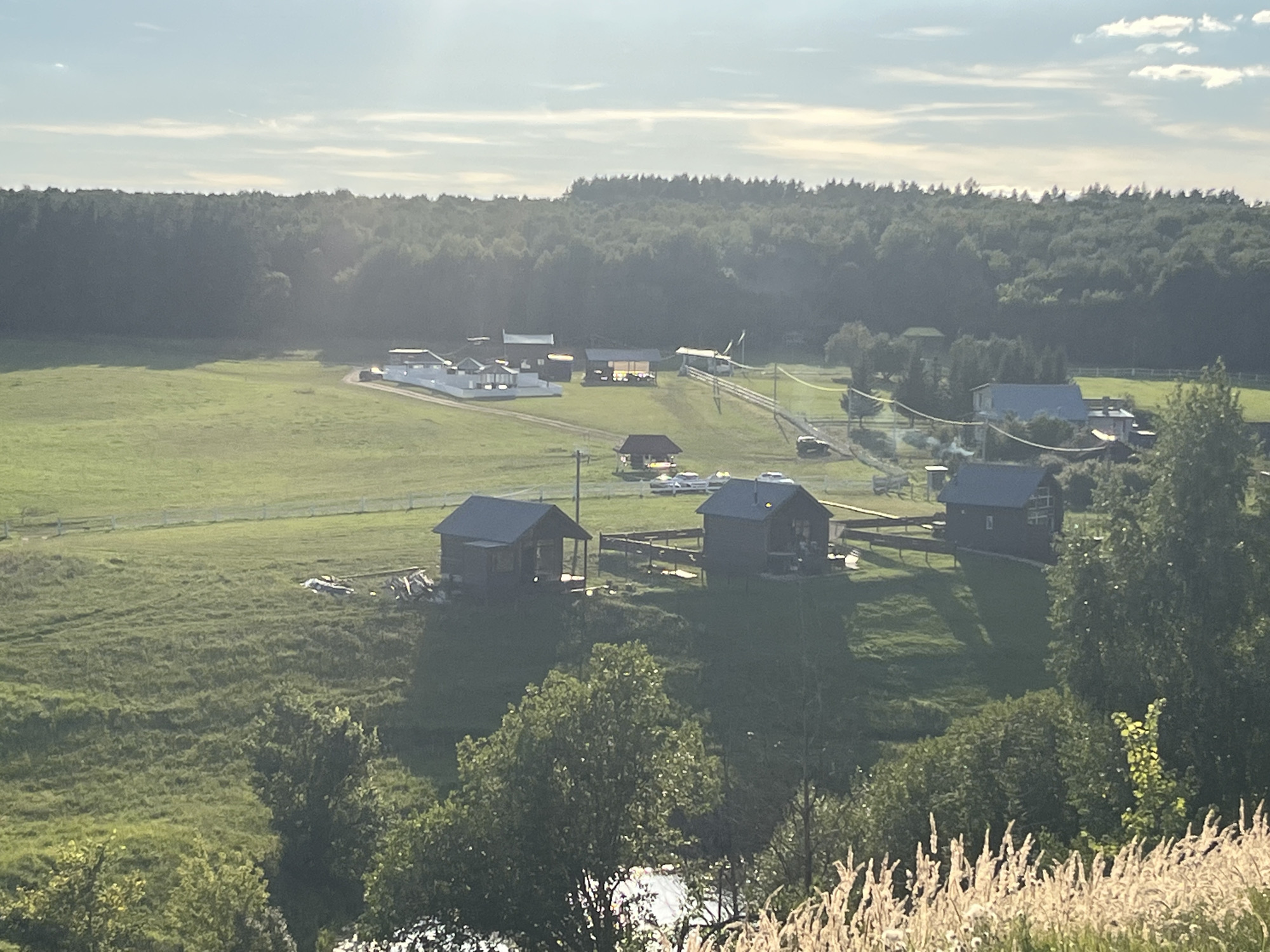 The image depicts a rural landscape in Srednie Pinyachi, Russia. The scene is characterized by rolling green hills covered in grass, dotted with trees and several buildings. In the foreground, dry, tall grasses are visible on a hill. Further into the scene, there are several wooden structures, some appearing to be houses with dark roofs and windows, situated on elevated ground. One cluster of buildings in the middle ground features white structures with dark roofs, possibly a residential complex or farm buildings, with a road leading to them. A dark forest borders the area in the background under a pale sky with scattered clouds. The sunlight suggests it is either early morning or late afternoon. There are no visible people in the image.