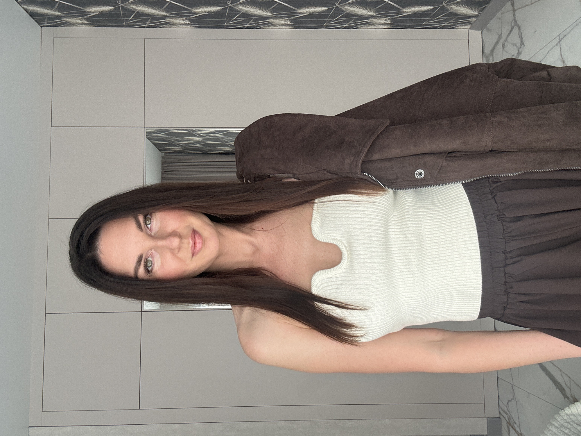 The scene is an indoor domestic setting in the city of Chaikovsky, Russia. A single adult female stands, facing the camera directly, from the waist up. She has long, straight, dark brown hair and green eyes, with light makeup visible. She wears a white, ribbed, strapless top featuring a V-shaped notch at the neckline, paired with a dark brown, possibly pleated, skirt with an elastic waistband. A brown suede or faux-suede jacket, with a visible silver zipper and a silver snap button on the chest, is draped over her left shoulder. The background consists of a light gray, floor-to-ceiling modular wardrobe or cabinet system, one panel of which includes a mirror reflecting a dark curtain and a portion of the adjacent wall. To the right, part of a wall is covered with dark wallpaper featuring a pattern of light-colored, abstract linear elements resembling feathery fronds. The visible floor is composed of white marble or marble-effect tiles with grey veining.