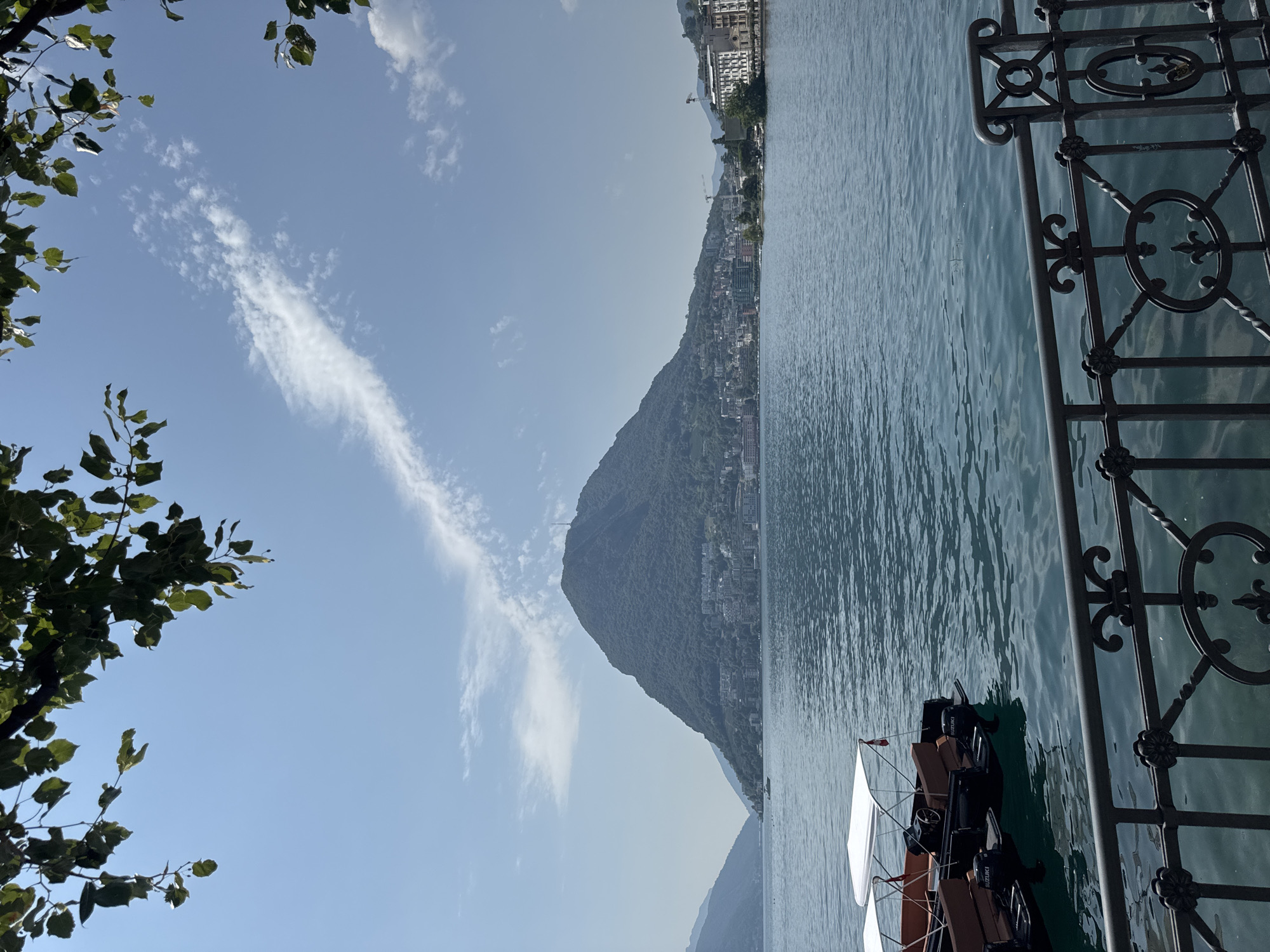 The image depicts a serene view of Lake Lugano in Lugano, Switzerland. In the foreground, a black metal fence with decorative elements runs along the bottom of the frame. Beyond the fence, two small boats with white canopies are moored on the calm, blue water, each with a "Suzuki" engine visible.
The middle ground is dominated by the expansive, shimmering surface of the lake. In the background, the imposing Monte San Salvatore mountain rises, its slopes covered in lush greenery and dotted with buildings that form the outskirts of the city of Lugano. A tall telecommunications tower is visible at the summit of the mountain. The city's architecture, characterized by a mix of modern and older buildings, lines the shoreline.
The sky is a clear, pale blue, with a single, wispy white cloud trail stretching diagonally across it. Green leaves and branches from an overhanging tree frame the top of the image, adding a natural element to the urban and natural landscape. The overall atmosphere is tranquil and picturesque, suggesting a bright, sunny day.
