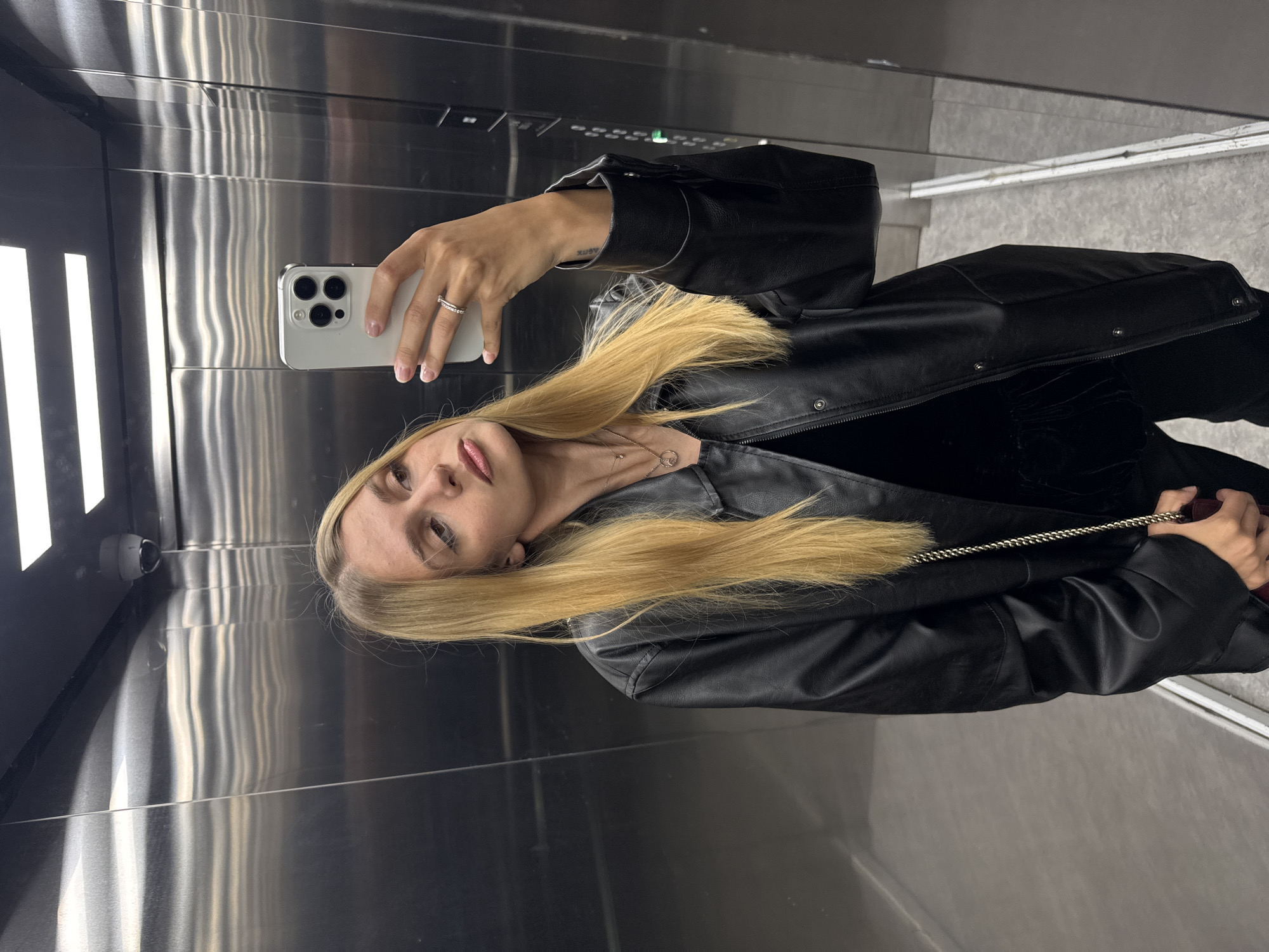 A woman is taking a selfie in an elevator. The elevator is likely in the city of Unknown, Unknown. She is holding a white smartphone in her right hand and posing for the camera. She has long, blonde hair and is wearing a black leather jacket. The elevator is made of stainless steel and has a security camera installed in the upper left corner. The floor of the elevator appears to be light gray.