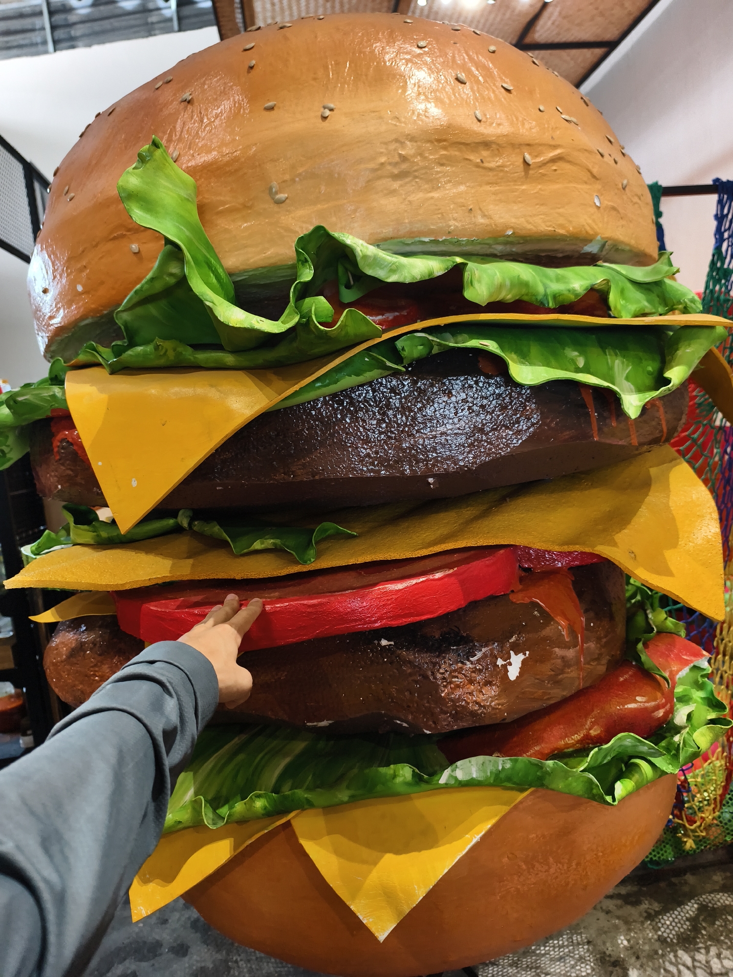The image shows a very large, multi-layered, model burger. A person's hand points to the red tomato layer of the burger. The burger is composed of layers including the top bun with sesame seeds, lettuce, tomato slices, a brown patty, and yellow cheese slices. The burger appears to be inside, possibly in a restaurant or attraction. There are other items in the background including some shelving and some colored netting. Based on the prompt instructions, it is located in Bac Nha Trang Ward, Vietnam.