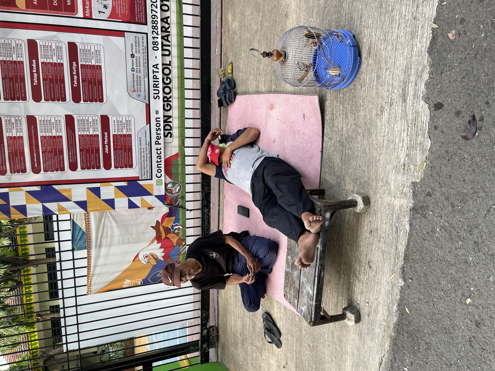 Two elderly Indonesian men are sitting on the ground outside a school. One man, wearing a dark t-shirt and blue pants, is seated cross-legged on a pink mat, holding what appears to be a lighter in his hand. Next to him, a flip-phone rests on the mat. The other man, wearing a dark t-shirt with a graphic and dark pants, is lying down on a similar mat, with his arm behind his head. A cage containing three birds sits on the ground in front of him. A pair of black flip-flops are on the ground to the left of the seated man, and a pair of black slides are near the lying man. The men are sitting near a low, rustic wooden bench supported by stones. Behind them, a fence separates the outdoor area from a building with trees and a sports court visible. A banner with colorful geometric patterns and an illustration of a bird is attached to the fence. Text on the banner indicates it is for "SDN GROGOL UTARA 01" and provides contact information for "SURIPTA - 0812889720". The pavement in front of them is made of concrete and asphalt, with some debris and leaves scattered on the ground. The scene appears to be in South Jakarta, Indonesia, based on the school's name and the presence of Indonesian text.