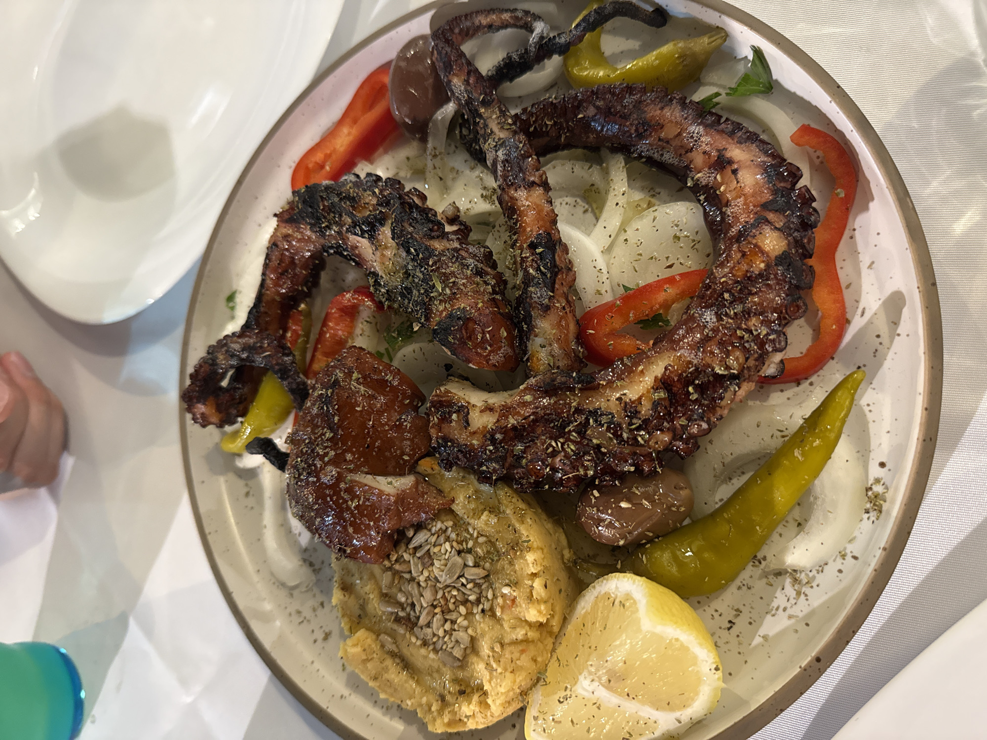 A top-down, medium shot captures a platter of grilled octopus served with assorted accompaniments on a white-clothed table, likely in Pallini Municipal Unit, Greece. The octopus tentacles, charred and glistening, are arranged in a circular pattern atop a bed of thinly sliced white onions. Red and yellow bell pepper strips, olives, and a wedge of lemon are strategically placed around the octopus. To the left of the platter, a mound of hummus, sprinkled with seeds, is visible, with a piece of what appears to be roasted tomato or meat adjacent to it. The plate itself is a light beige with a darker rim, resting on a white tablecloth. In the background, out of focus, a bright turquoise beverage in a glass and a portion of another white plate are present. The lighting is bright, suggesting an indoor or covered outdoor dining setting.