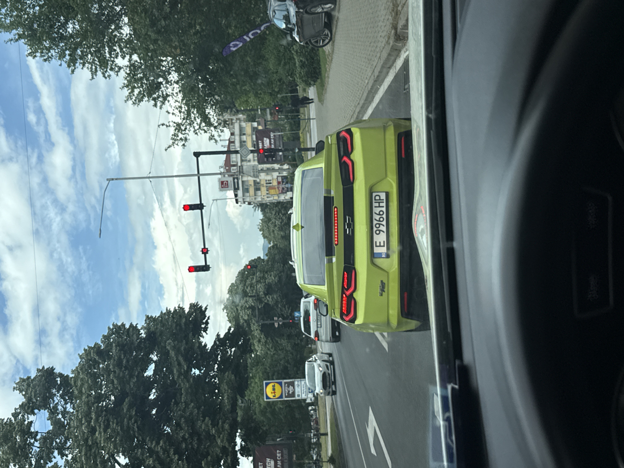 The image captures a street scene in Blagoevgrad, Bulgaria. It is taken from the interior of a vehicle, looking out at traffic stopped at a red light. The most prominent object is a bright lime-green Chevy Camaro with a Bulgarian license plate "E 9966 HP". The car is positioned directly ahead in the same lane. Behind the Camaro, other vehicles are also waiting at the intersection, including a white van and a smaller white car. The street is lined with trees, traffic lights, and commercial signs, including a Lidl supermarket sign. Buildings are visible further down the street. The sky is partly cloudy.