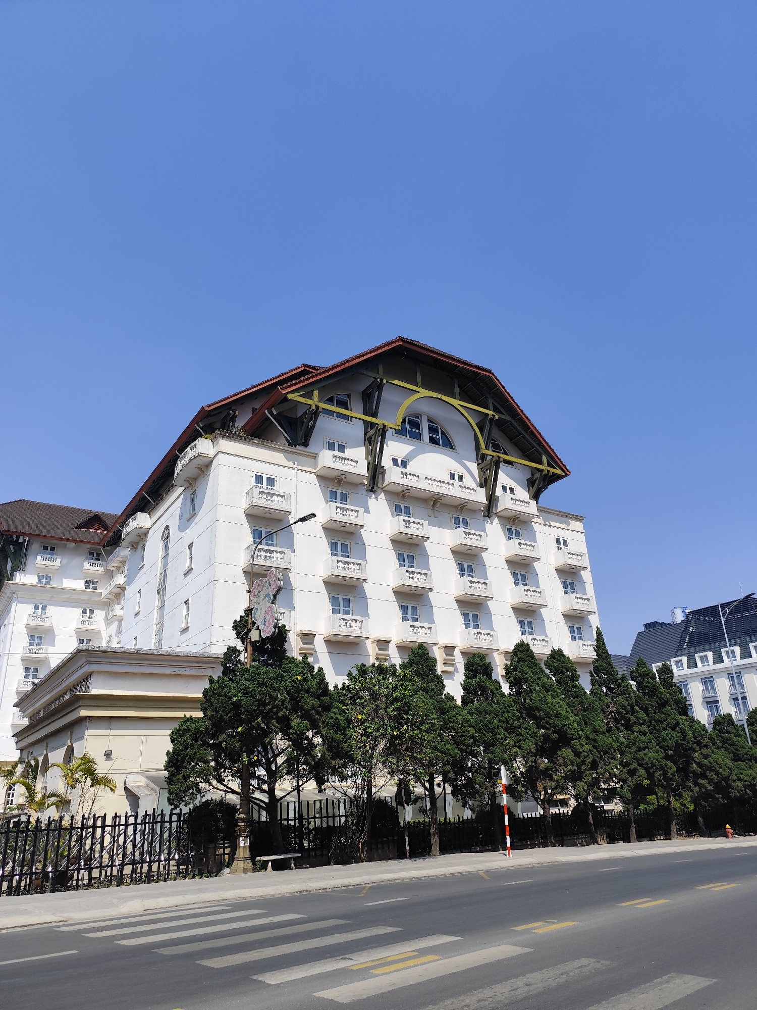 The image depicts a low-angle shot of the Hotel du Parc Dalat, a historic hotel in Da Lat, Vietnam. The hotel is a prominent white building with multiple floors, each featuring small balconies. The roof has a distinctive red-tiled design and a unique arched structure with yellow supports. The building is partially obscured by a row of evergreen trees that line the street. A black wrought-iron fence runs along the property line. A street with yellow lane markings and a pedestrian crosswalk is visible in the foreground. The sky is clear and blue. Another building with a dark roof can be seen in the background to the right of the hotel.