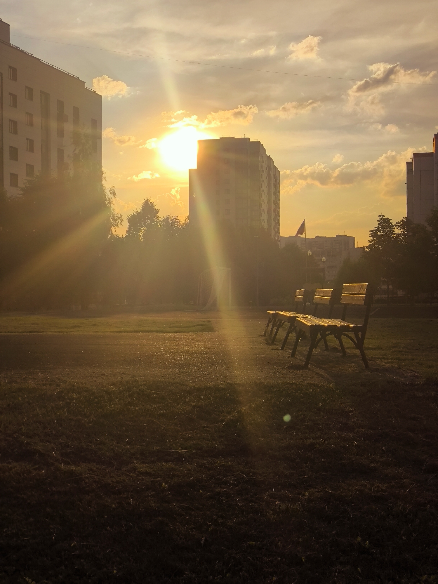 In a city park at sunset, three empty wooden benches sit on a grassy area, facing away from the camera towards a large sunbeam. The sun, partially obscured by clouds, casts a warm golden light across the scene, creating lens flare and highlighting the dust motes in the air. In the background, apartment buildings and trees are silhouetted against the bright sky. A flag flies from a pole on one of the buildings. The foreground is dark, with the grass and ground dimly lit by the fading sunlight.