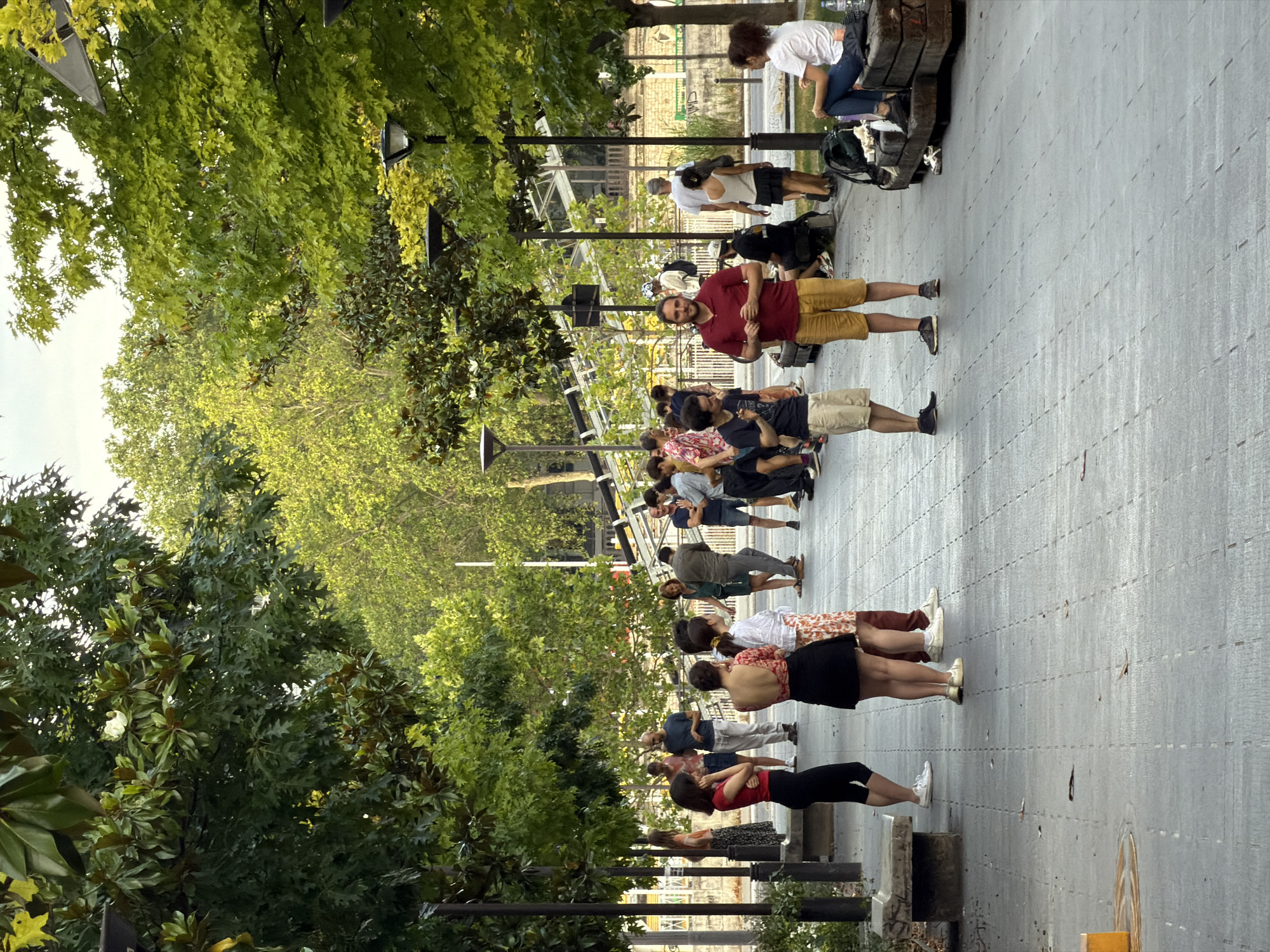 The image depicts a group of people gathered on a paved walkway lined with trees. The setting appears to be a public space, possibly a park or a promenade. Several individuals are standing in conversation, while others are seated on benches. Some people are casually dressed, suggesting a warm day. The trees are lush and green, indicating a temperate season. The background shows architectural elements and more greenery, suggesting an urban environment. While there are no explicit indicators within the image itself, the context of a Parisian promenade with trees and public gathering spaces suggests this could be a location like the Coulée Verte René-Dumont or a similar tree-lined avenue in Paris, France.
