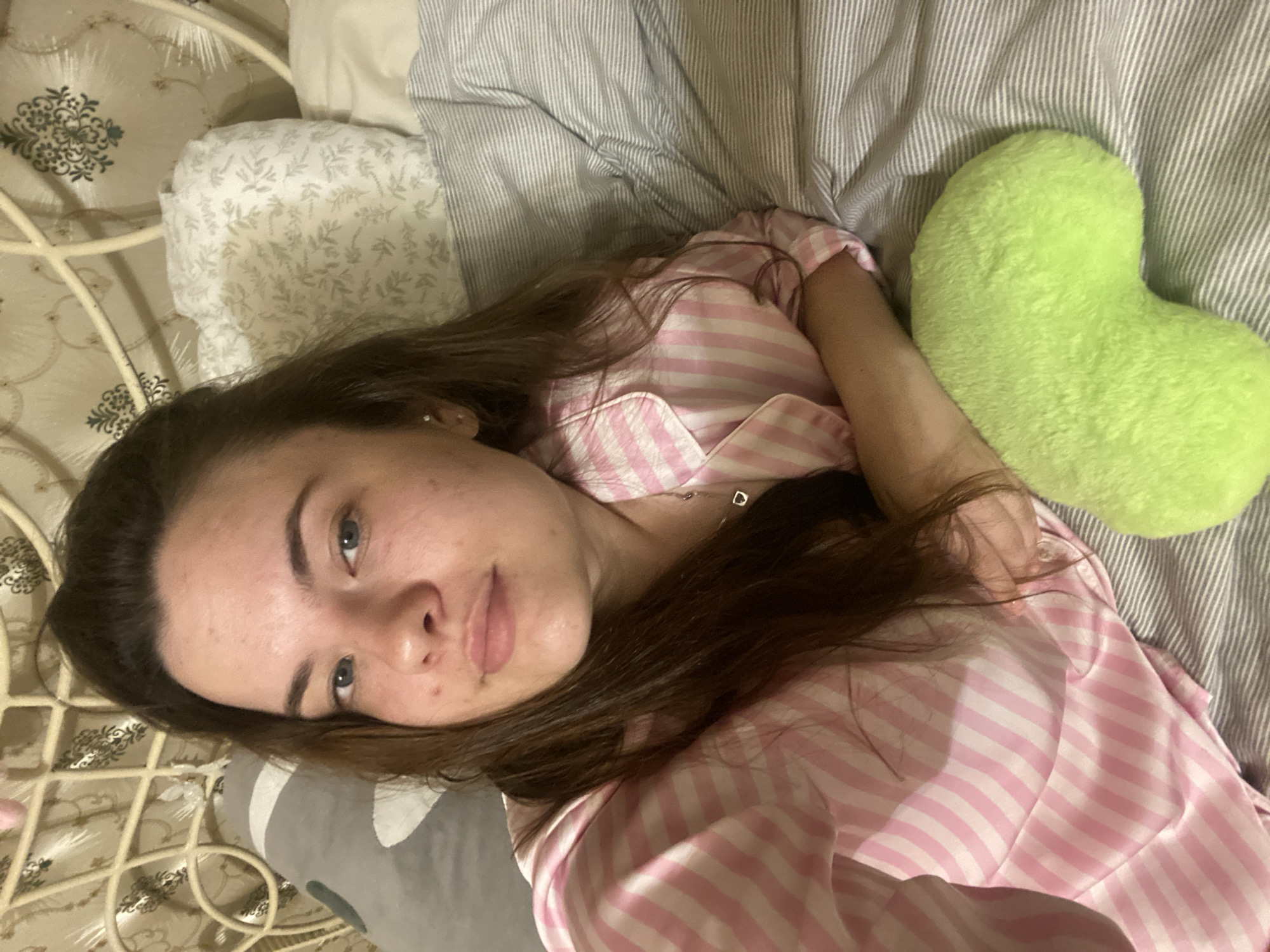 A young woman with long brown hair, wearing pink and white striped pajamas, is lying in bed. She is looking at the camera with a neutral expression. The bed is made with striped gray and white sheets. Behind her, there are several pillows, one of which is gray and has a sloth pattern, and another is white with a floral pattern. A cream-colored metal headboard is visible in the background, adorned with decorative scrollwork. To the right of the woman, there is a bright green, heart-shaped pillow. The scene appears to be an indoor bedroom environment. The image does not provide any specific indicators for its location within the city of Poltava, Ukraine.