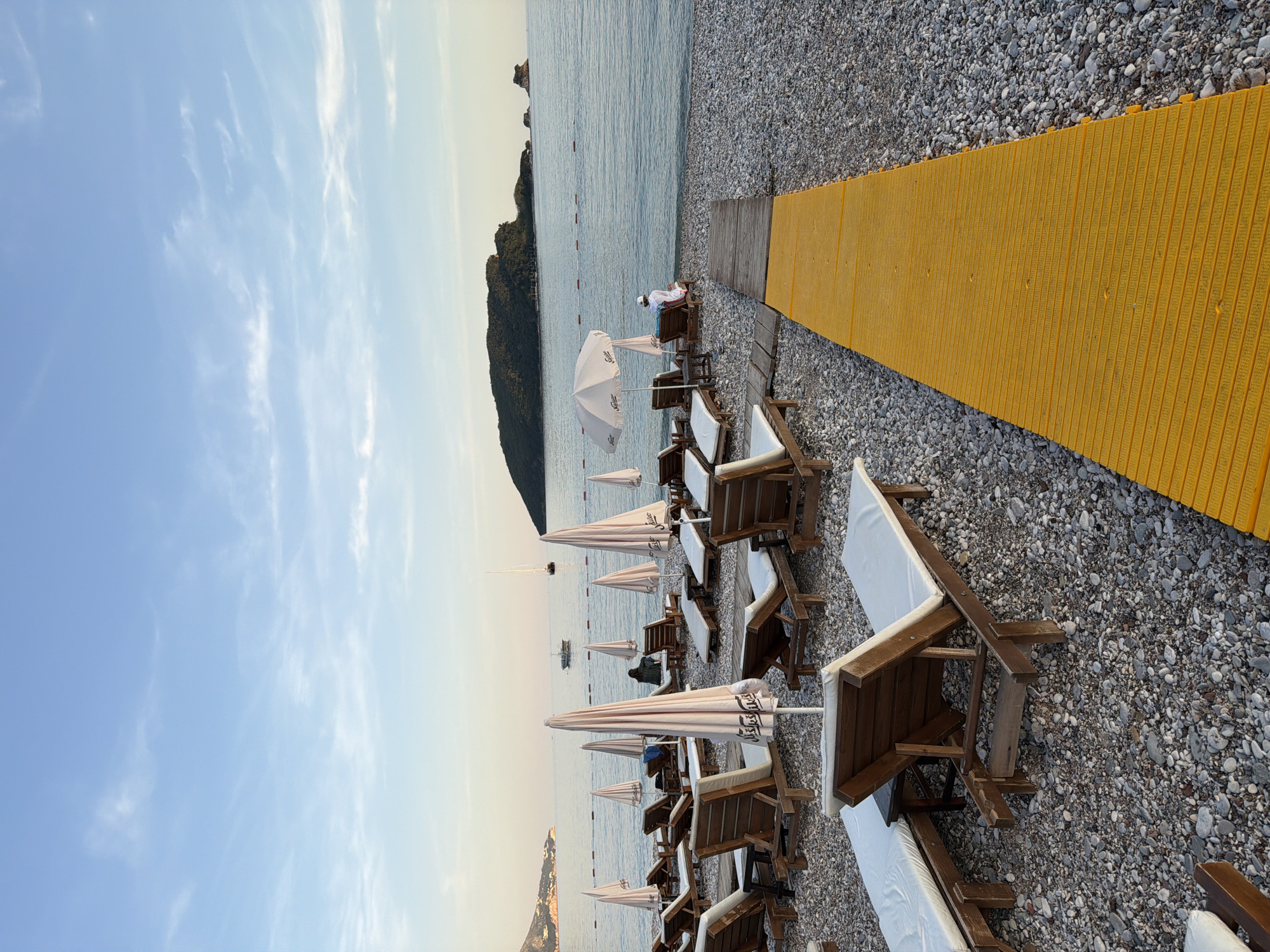The image shows a pebble beach in Budva, Montenegro. The beach is lined with wooden beach chairs, most of which have white cushions, and white umbrellas. A yellow walkway extends from the foreground toward the sea, transitioning to a section of wooden boards before reaching the water. In the background, there is a verdant, rocky island. A few boats are visible on the water, and the sky is light blue with scattered clouds. Some people are seated on the chairs near the end of the wooden walkway.