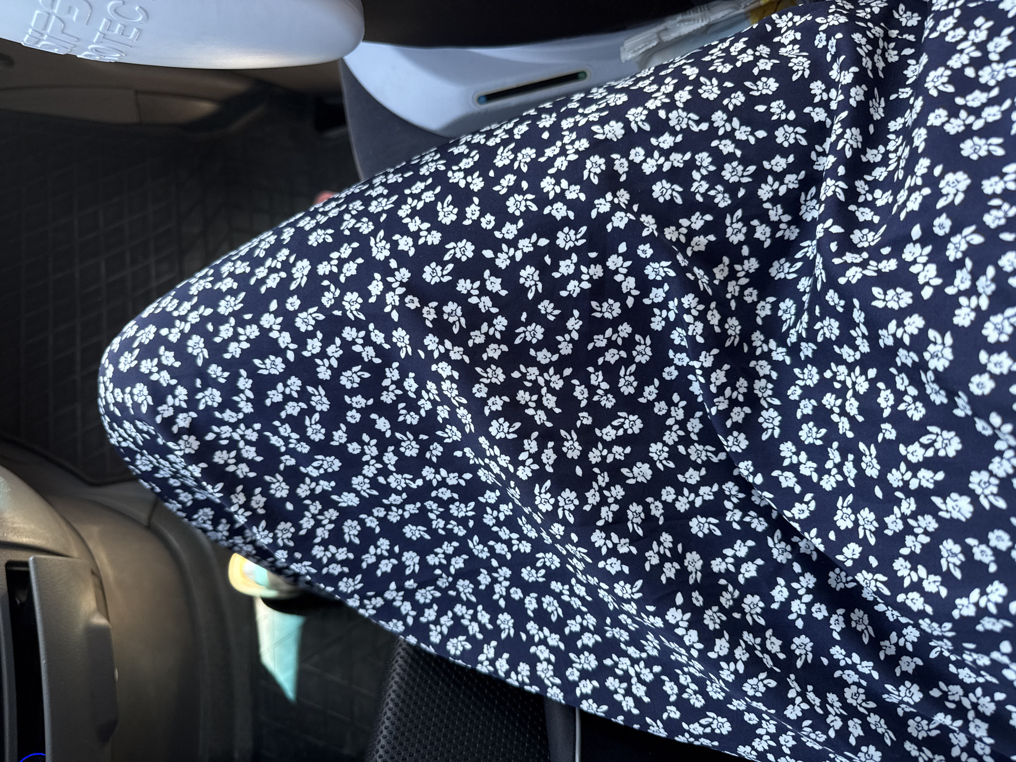 The image shows a close-up view of a person's legs wearing a dark blue dress with a white floral pattern. The person is sitting in a vehicle, as indicated by the car interior visible in the background, including a gray dashboard and black floor mats. Sunlight is entering the car from the left, casting a shadow on the floor. To the right, a portion of a white object, possibly a car seat or a container, is visible with text that appears to say "SIPS PROTEC." The image does not provide any specific information to locate it in Volpertshausen, Germany.