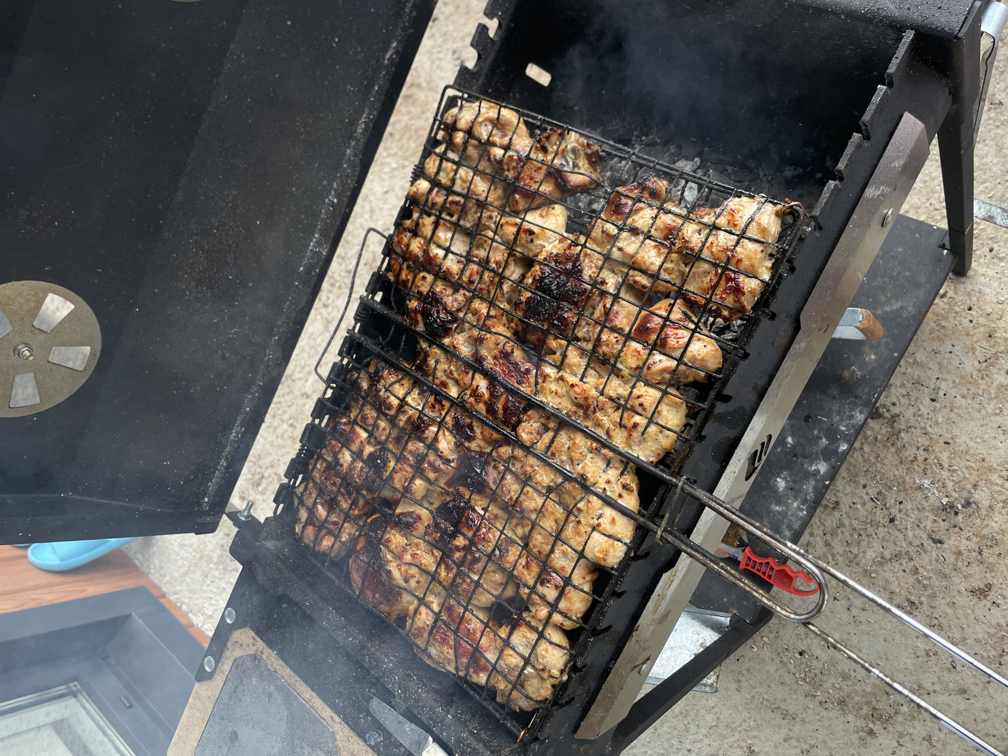 The image depicts a grill with food cooking on it. It appears to be some type of cubed meat, possibly chicken, in a wire basket. The grill itself is black and shows signs of use. There's some smoke coming from the grill, indicating the food is actively being cooked. The surrounding environment suggests the grill is situated outdoors on a concrete surface. No people are visible in the frame. The location, Подшивалово, Russia, is provided, but there are no visual cues in the image to independently confirm this location.