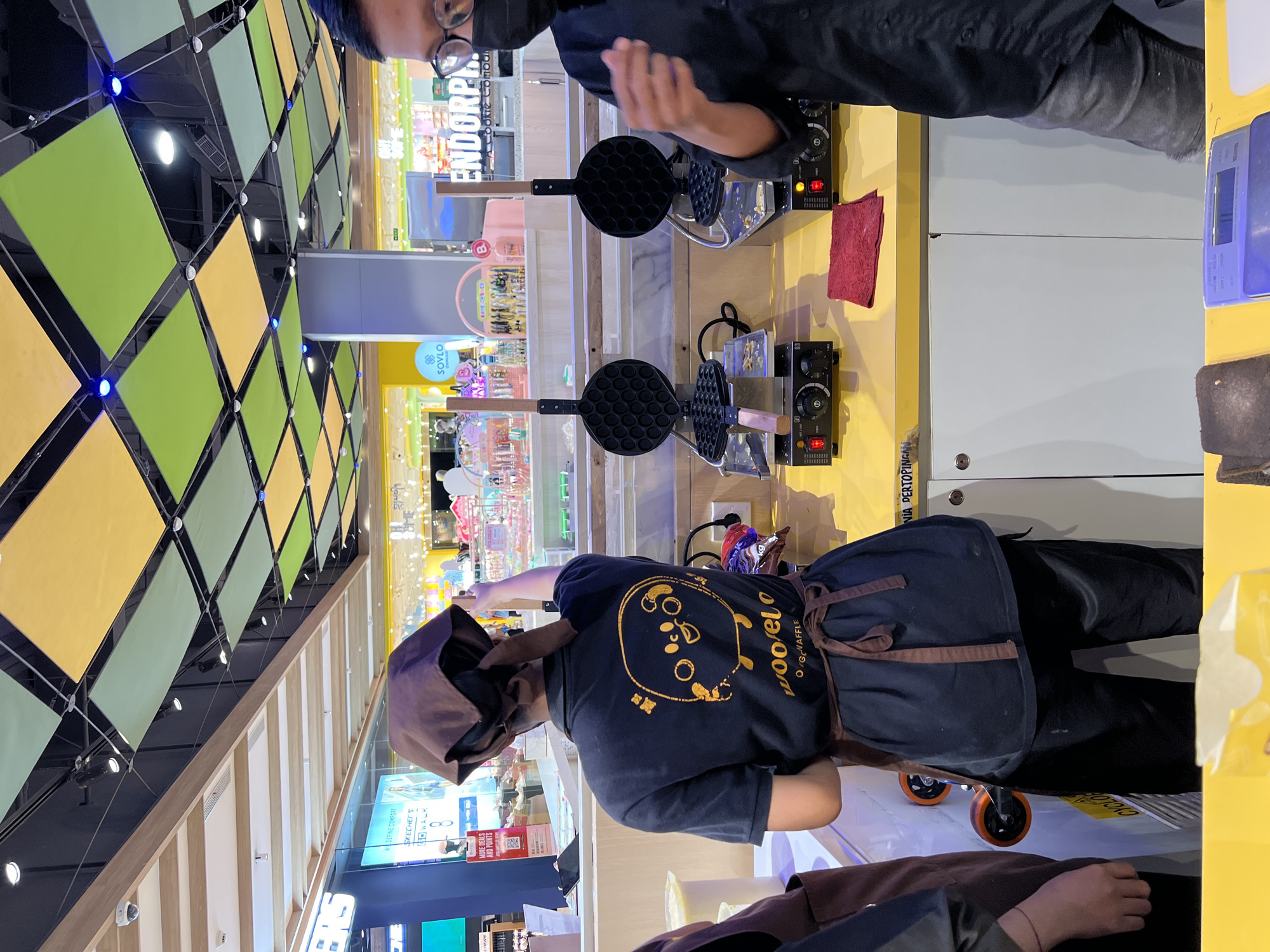 In Denpasar, Indonesia, a person wearing a brown apron over a dark t-shirt with a cartoon waffle character printed on the back stands behind a yellow counter. They are wearing a brown headscarf that covers their hair. In front of them are two waffle makers, each with a black, round, hexagonal-patterned mold, and a wooden handle. One waffle maker has a small amount of batter visible in its mold. A red cloth is placed on the counter to the right of the waffle makers. To the right of the counter, another person wearing glasses, a black face mask, and a black shirt is partially visible. In the background, there are shops with brightly colored displays, including a sign that reads "ENDORPHIN." The ceiling features a grid of green and yellow square panels with blue lights.