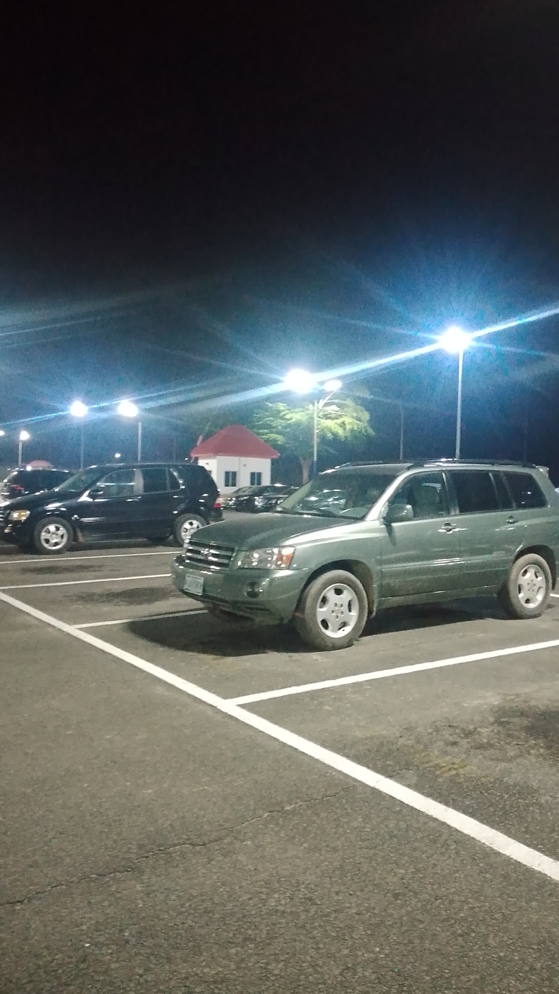 The photograph was taken at night in a parking lot in Orerokpe, Nigeria. The lot is well-lit by overhead streetlights. In the foreground, a green SUV is parked within painted white lines, designating a parking space. Behind it, to the left, a dark-colored SUV is parked in a similar fashion. Further back, a small, white building with a red roof is visible. Other cars are parked nearby, though details are obscured by distance and lighting. The overall environment suggests an urban or commercial area.