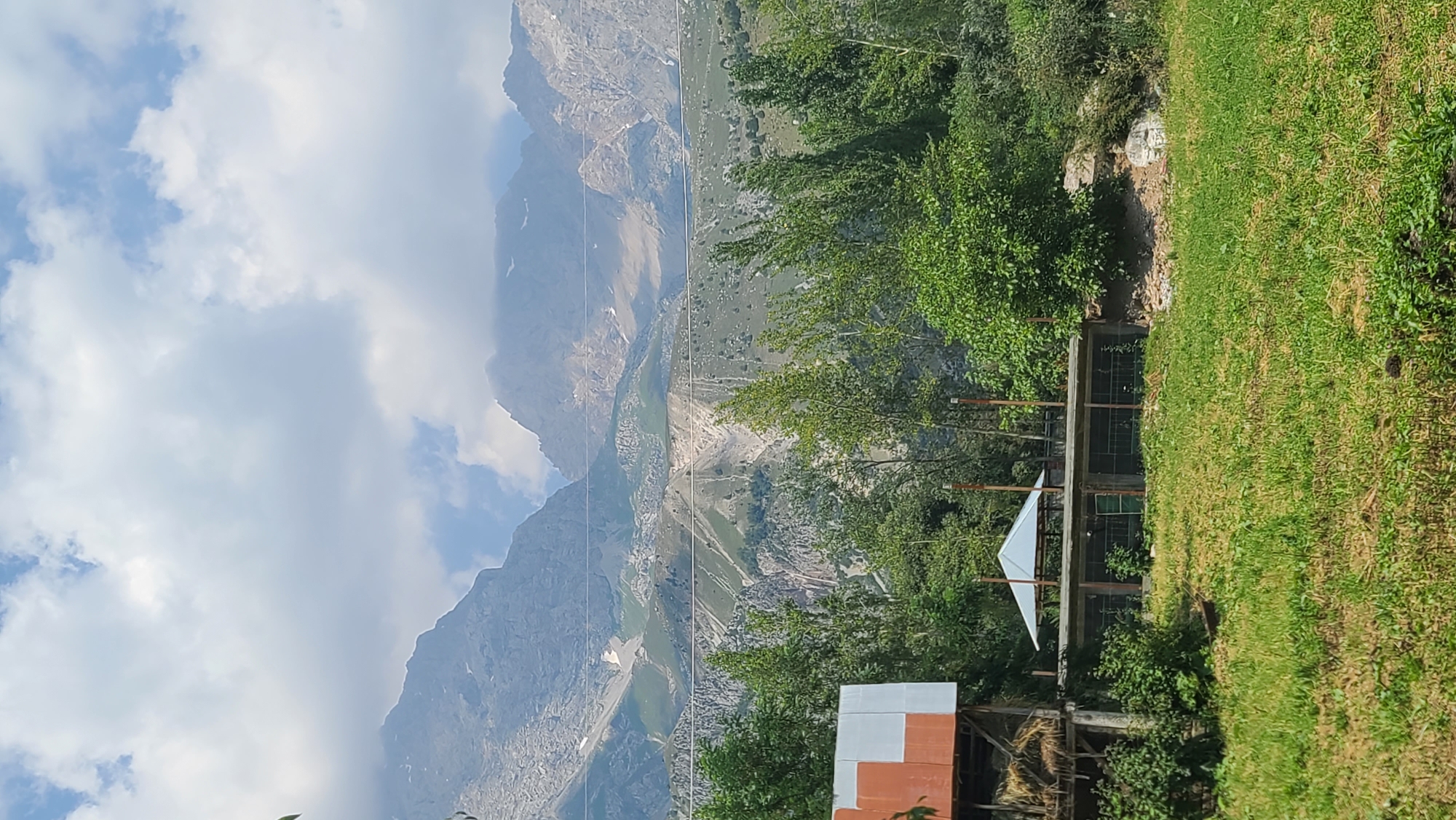 The image depicts a mountainous landscape in Arstanbap, Kyrgyzstan. In the foreground, a grassy field leads to a series of structures. To the left, a part of a building with a red and white corrugated metal roof is visible, with hay bales stacked beneath it. Adjacent to this, a long, low structure with an open roof section covered by a white canopy extends horizontally. This structure appears to be an enclosure, possibly for animals, with wire mesh sides and wooden posts.
Behind these structures, a dense line of green trees forms a natural barrier. Beyond the trees, the terrain rises dramatically, revealing steep, rocky mountainsides with patches of green vegetation and snow-capped peaks under a cloudy sky. Power lines are visible crisscrossing the middle ground, stretching across the mountains. The overall scene suggests a rural setting nestled within a grand natural environment. No people are visible in the image.