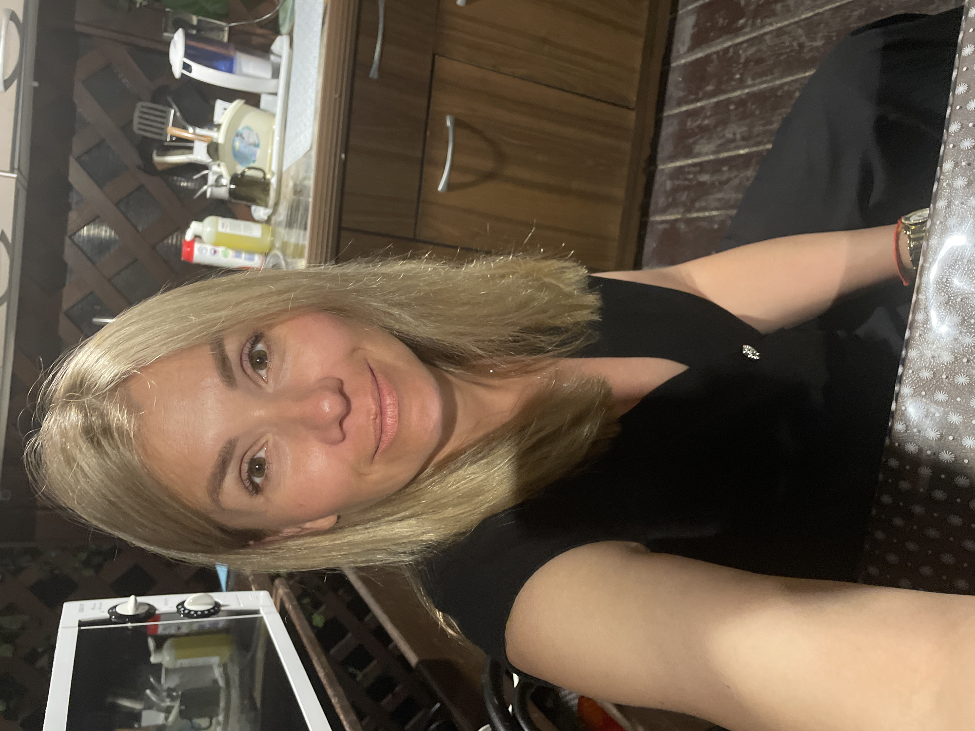 A woman with blonde hair, wearing a black dress and a watch, takes a selfie. She is seated in what appears to be an outdoor kitchen area, with wooden floors and cabinets visible. A microwave is present on a shelf to her left, and various kitchen items are on the counter behind her. The lighting suggests it is evening or indoors.