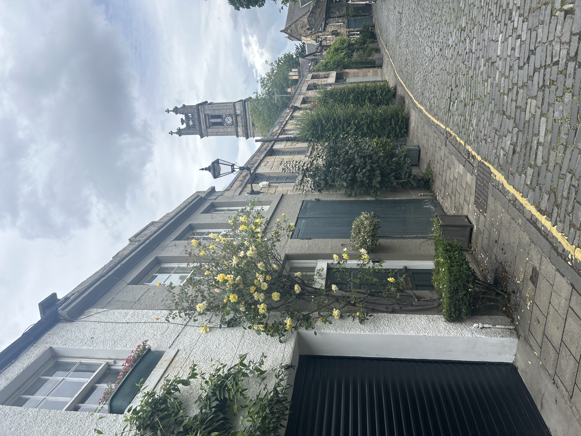 This media file captures a view of a charming street in Edinburgh, United Kingdom, characterized by historic stone buildings and a cobblestone pathway. In the foreground, a light-colored, textured building occupies the lower left, with a large, dark garage door. Above the garage door, a window is adorned with a planter overflowing with pink flowers. Further up the building, a climbing vine with yellow roses is in bloom, extending across the facade and framing another window. The building's architecture features contrasting textures, with a rougher lower section and a smoother upper portion.
To the right of the foreground building, a cobblestone street is visible, marked by a bright yellow line running along its edge. The street is lined with more stone buildings and lush green bushes. In the middle ground, a prominent clock tower rises above the buildings, featuring a clock face and decorative architectural elements. The tower suggests the presence of a church or significant public building.
The scene is set under a cloudy sky, indicating overcast weather. The lighting is diffused, and there are no strong shadows, suggesting it is daytime, likely mid-morning or afternoon. The overall atmosphere is one of historical charm and quiet urban life. No people are visible in the frame, focusing attention on the architecture and natural elements. The presence of the clock tower and the distinctive stone construction strongly points to Edinburgh as the location. The image is oriented vertically.
