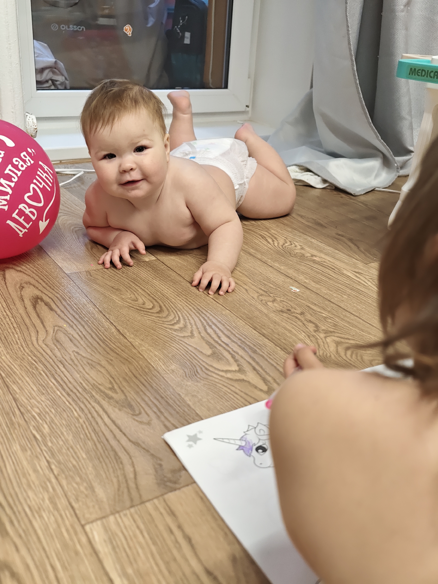 A fair-skinned infant wearing a diaper lies on a light brown wood-grain floor. The infant is in a prone position, head up, and looking at the camera. Next to the infant is a pink balloon with Russian text on it. In the foreground, a child is drawing a unicorn on a sheet of white paper. The scene is indoors, near a window with "OLSSON" printed on it and a medical stand. Given the Russian text on the balloon, the location is likely Chaikovsky, Russia, as requested.