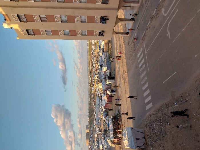 An aerial view shows a settlement next to a paved road. Buildings border one side of the road, while tents and structures form the settlement on the other. Pedestrians walk alongside the road and cross at a marked crosswalk. The sky is blue with scattered clouds. The scene suggests a densely populated area possibly near a building.