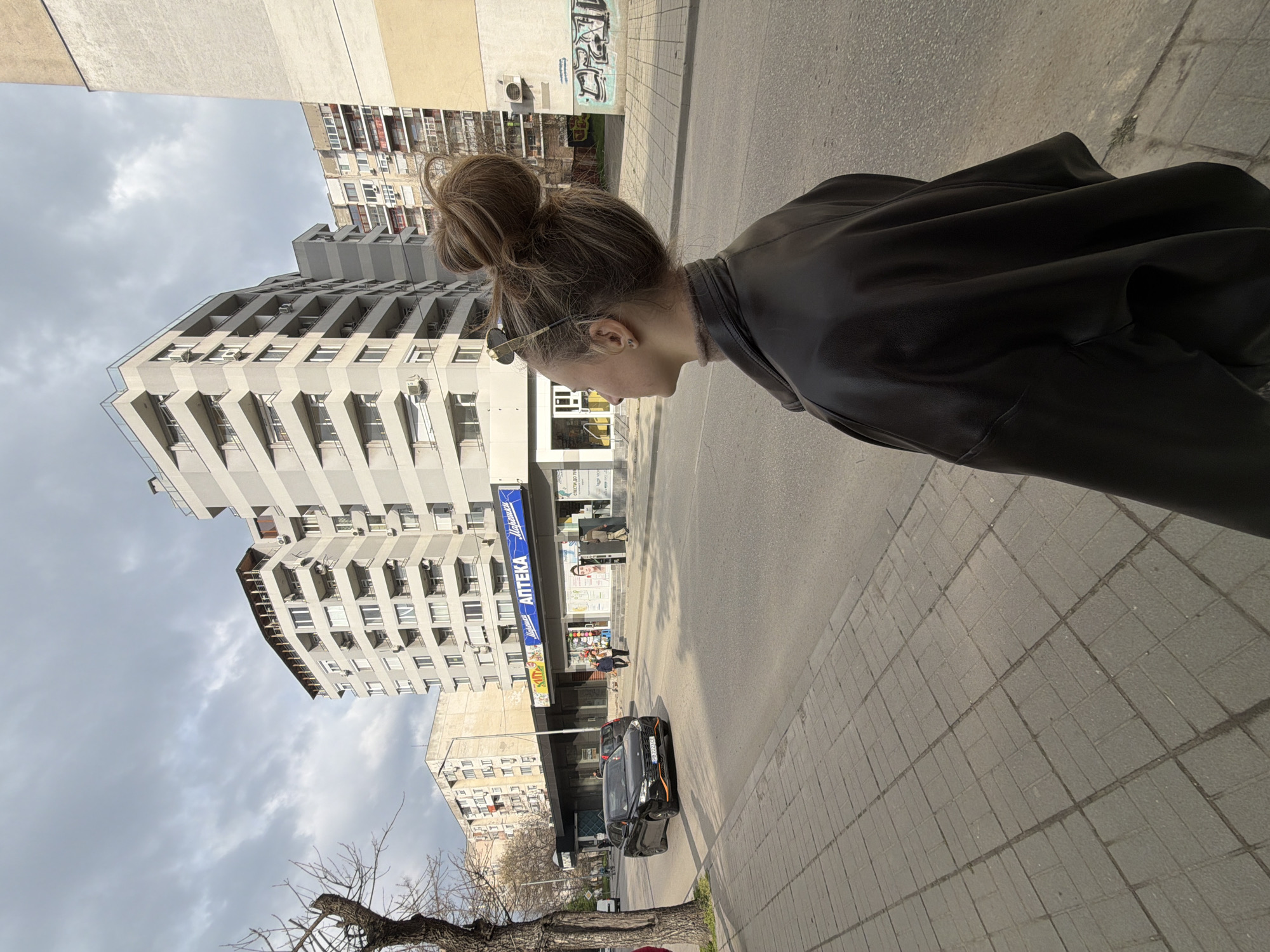 The image captures an urban street scene in Plovdiv, Bulgaria, specifically looking southeast along Bulevard Vasil Aprilov near the intersection with ulitsa Vaska Nedyalkova. In the immediate foreground, a woman is seen from her left mid-back, standing on a grey rectangular paved sidewalk. She has brown hair styled in a high bun, with sunglasses perched on top, and wears a dark, possibly leather, jacket over a brown turtleneck.
Across a two-lane asphalt road, a black BYD Atto 3 SUV, distinguished by orange accents and bearing Bulgarian license plate PB 2223 CG, is visible in the left lane. Behind it, another car is partially obscured. On the opposite side of the street, a multi-story light-grey residential building dominates the midground. Its ground floor houses a commercial establishment with a prominent blue sign displaying "АПТЕКА" (Pharmacy) in Cyrillic, accompanied by "Нарешки." Two individuals are observed near the pharmacy entrance.
Further into the background, a cluster of various apartment buildings, some with balconies and external air conditioning units, line the street. A beige wall to the right of the pharmacy features visible graffiti. To the far left in the foreground, a bare-branched deciduous tree stands on the sidewalk. The sky is overcast with grey clouds, suggesting daylight conditions.
