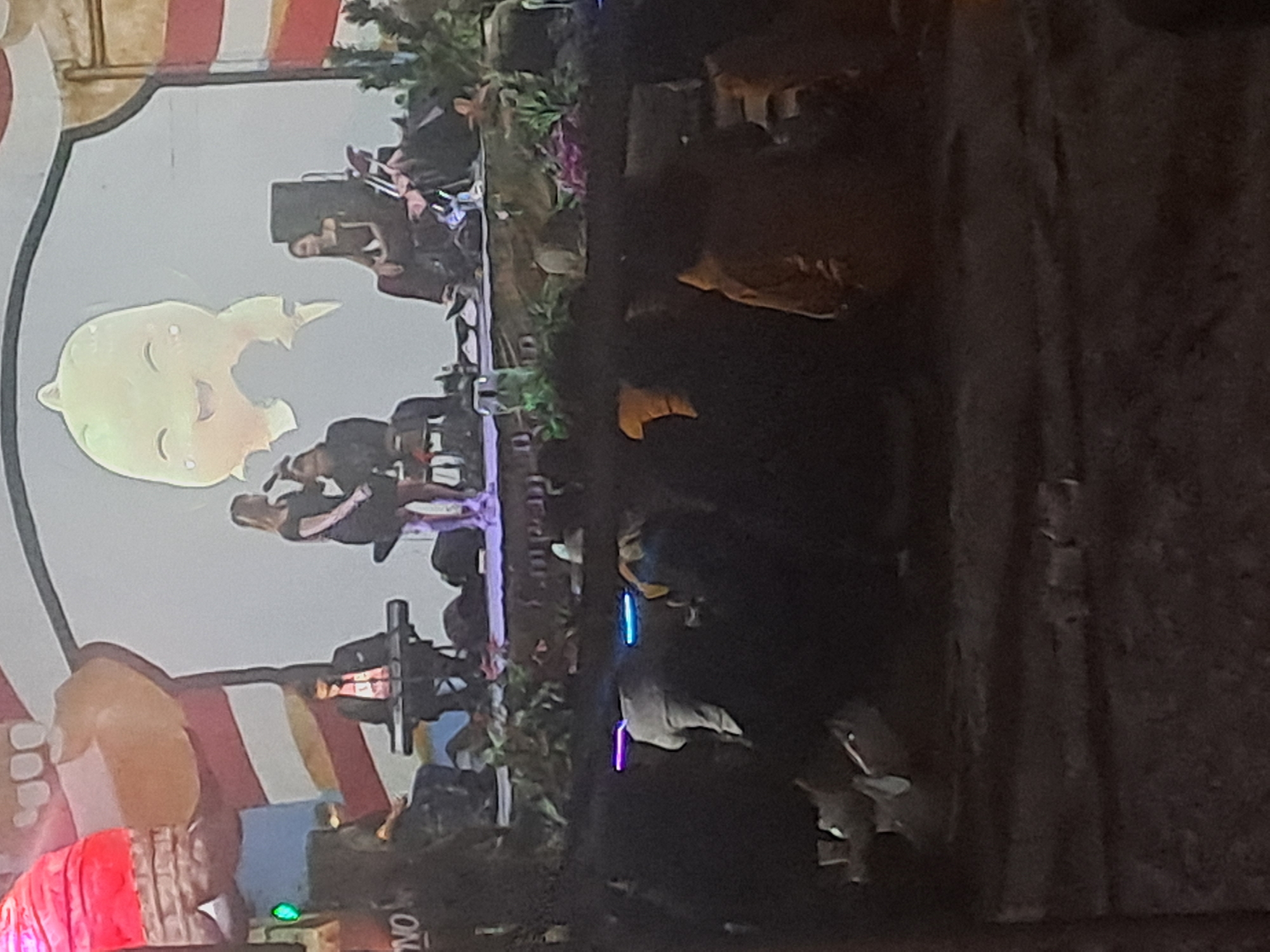 The image depicts an outdoor performance in Tirtomartani, Indonesia. A stage is set up with a backdrop featuring a cartoonish, yellow, smiling creature. On stage, a woman in a black dress stands holding a microphone, while another person plays a keyboard. A third person is seated behind the keyboard player. The stage is adorned with greenery. In front of the stage, an audience is seated on the ground, some with glowing lights. The overall atmosphere suggests a community event or concert.