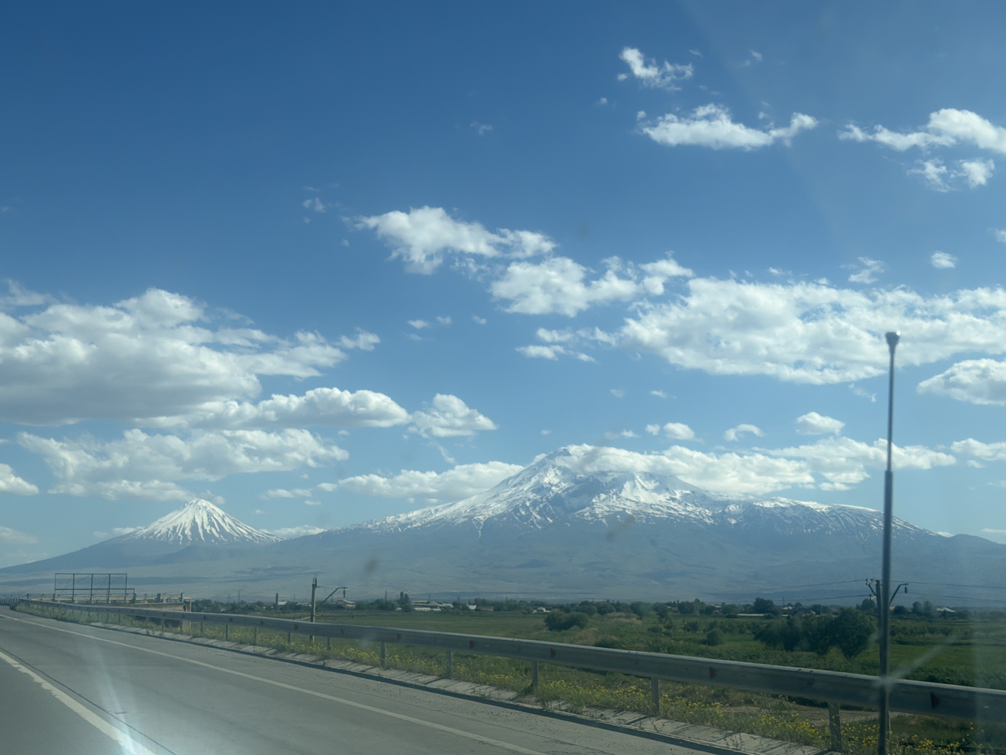 The image depicts a road leading towards two mountains, with the larger one partially covered in snow. A grey guardrail runs along the roadside. The sky is blue with scattered white clouds. A utility pole is visible on the right side of the image. The location is Unknown, Unknown.