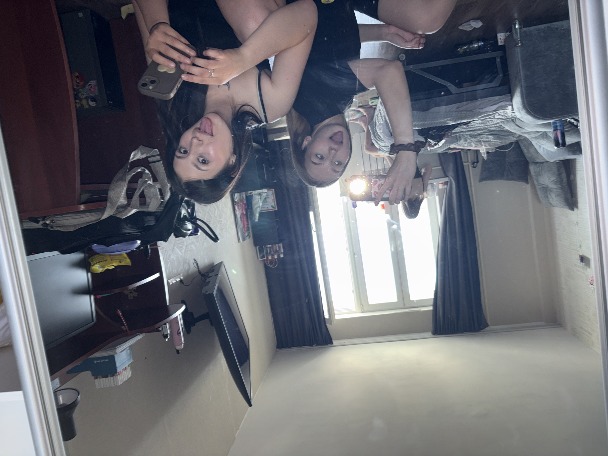 The image is a mirror selfie of two young women, seemingly taken in a room. One woman is on the left, and the other is on the right. Both are sticking their tongues out playfully. The environment shows a bedroom with a bed, curtains, and a TV. Based on the details, it can be identified as the city of Chaikovsky, Russia.