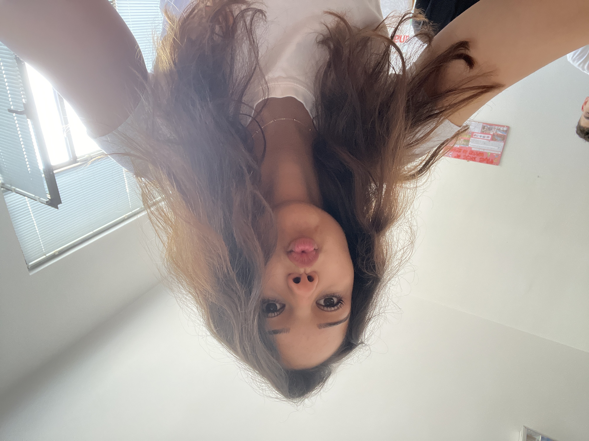 The image is an inverted selfie taken from a low-angle perspective, showing a young woman and the ceiling of an indoor room. The main subject is a woman with long, flowing brown hair, wearing a white t-shirt and a delicate silver-toned necklace around her neck. Her face is upside down, with wide-open eyes looking directly at the camera and her lips pursed in a "kissy face" expression.
The setting is a room with white walls and a white ceiling, brightly illuminated by natural light. On the left side, a window with horizontal blinds is visible, allowing bright sunlight to enter and create glare. A small portion of another person's head with short dark hair is visible in the upper right corner of the frame. On a wall in the upper right quadrant, there is a red rectangular object, possibly a calendar or poster, with some indistinguishable imagery and partially visible text, including the letters "eu". A small, light-colored object is barely visible in the lower right edge of the frame. The overall impression is a casual and playful moment, captured during the daytime.