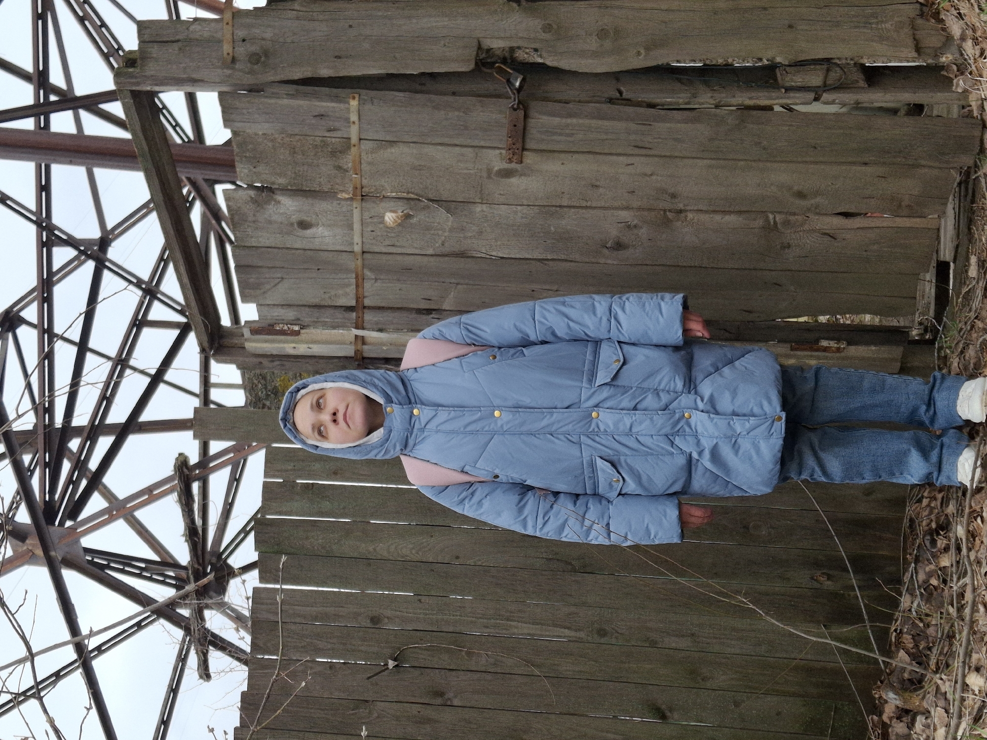 A vertical full-body shot captures a young person, likely a girl or young woman, standing outdoors. She is dressed in a light blue hooded puffer jacket with a contrasting pink inner lining visible around the hood, blue denim jeans, and white sneakers. Her head is tilted slightly upwards, and she looks towards the camera with a neutral expression.
The background is dominated by an old, weathered wooden fence or wall constructed from horizontal planks, displaying signs of age and decay, including cracks and discolored patches. A rusty padlock and chain are visible attached to one of the upper planks. Behind the wooden structure, on the left side of the frame, a large, intricate skeletal metal structure, possibly the remains of a disused industrial building or a water tower, rises against an overcast, bright sky. This metal framework consists of numerous dark, slender beams forming a complex geometric pattern.
The ground beneath the person's feet and at the base of the fence is covered with dry leaves, bare branches, and twigs, indicating a colder season such as late autumn, winter, or early spring. The person's warm attire further supports the cool weather inference. The overall scene suggests a desolate or neglected area, possibly an abandoned site in Chaikovsky, Russia. There is no visible text in the image.