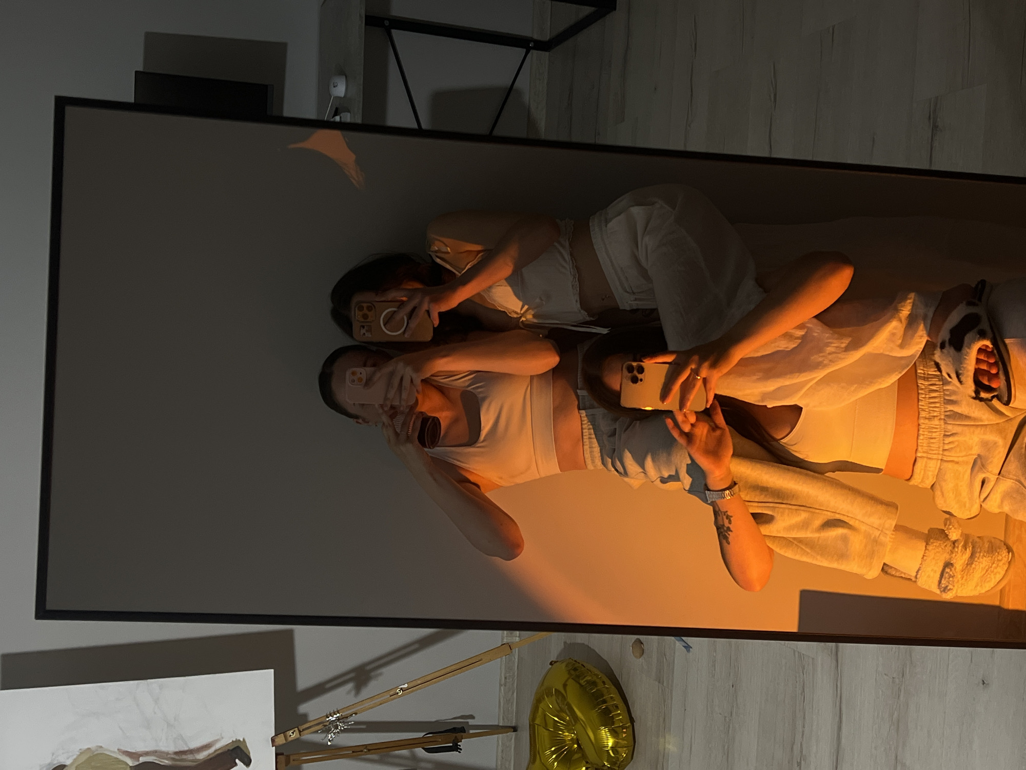 Three young women are taking a mirror selfie in a room. The woman in the foreground is sitting, facing away from the mirror, with her head tilted back and her arm raised to take the photo. She is wearing a white crop top and gray sweatpants with fuzzy slippers. The woman in the middle is sitting on the first woman's lap, facing the mirror and taking a photo. She is wearing a white crop top and gray sweatpants. The woman in the back is standing behind the middle woman, facing the mirror and taking a photo. She is wearing a white crop top and white pants. All three women are holding smartphones. A large, gold, heart-shaped balloon is visible to the left of the mirror. The room has light wooden floors and a large mirror. A desk and a computer monitor are visible in the background, suggesting the room may be a home office or bedroom. The scene is lit with warm, orange light, possibly from a lamp or sunset. The image does not contain any visual information that places it specifically in the city of Ternopil, Ukraine.
