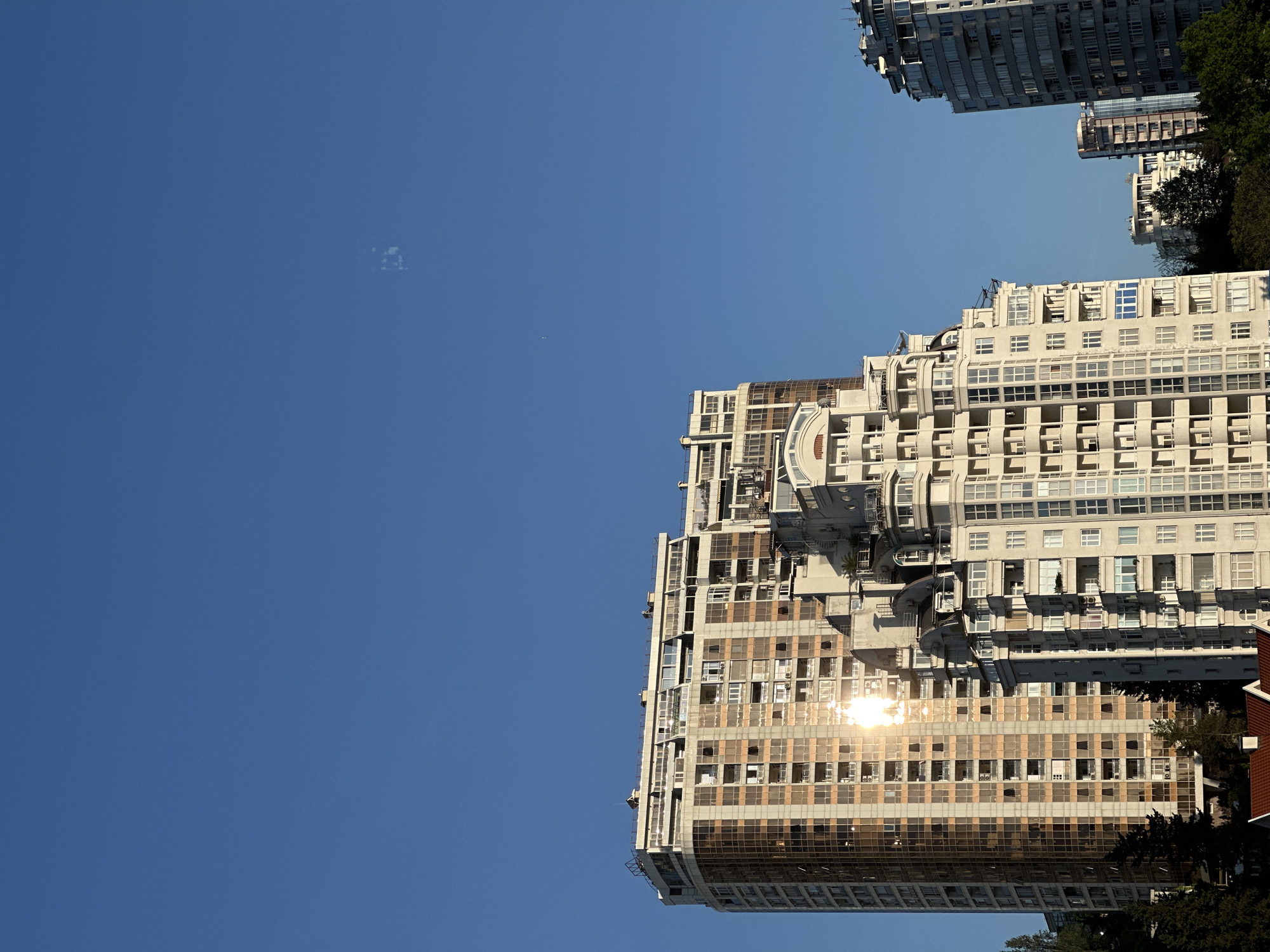 The image shows a tall, multi-story building with numerous windows in Sochi, Russia. The building appears to be modern residential or commercial architecture. Sunlight reflects brightly off some of the windows. The sky is clear blue, with some clouds. Other buildings and trees are visible in the background.