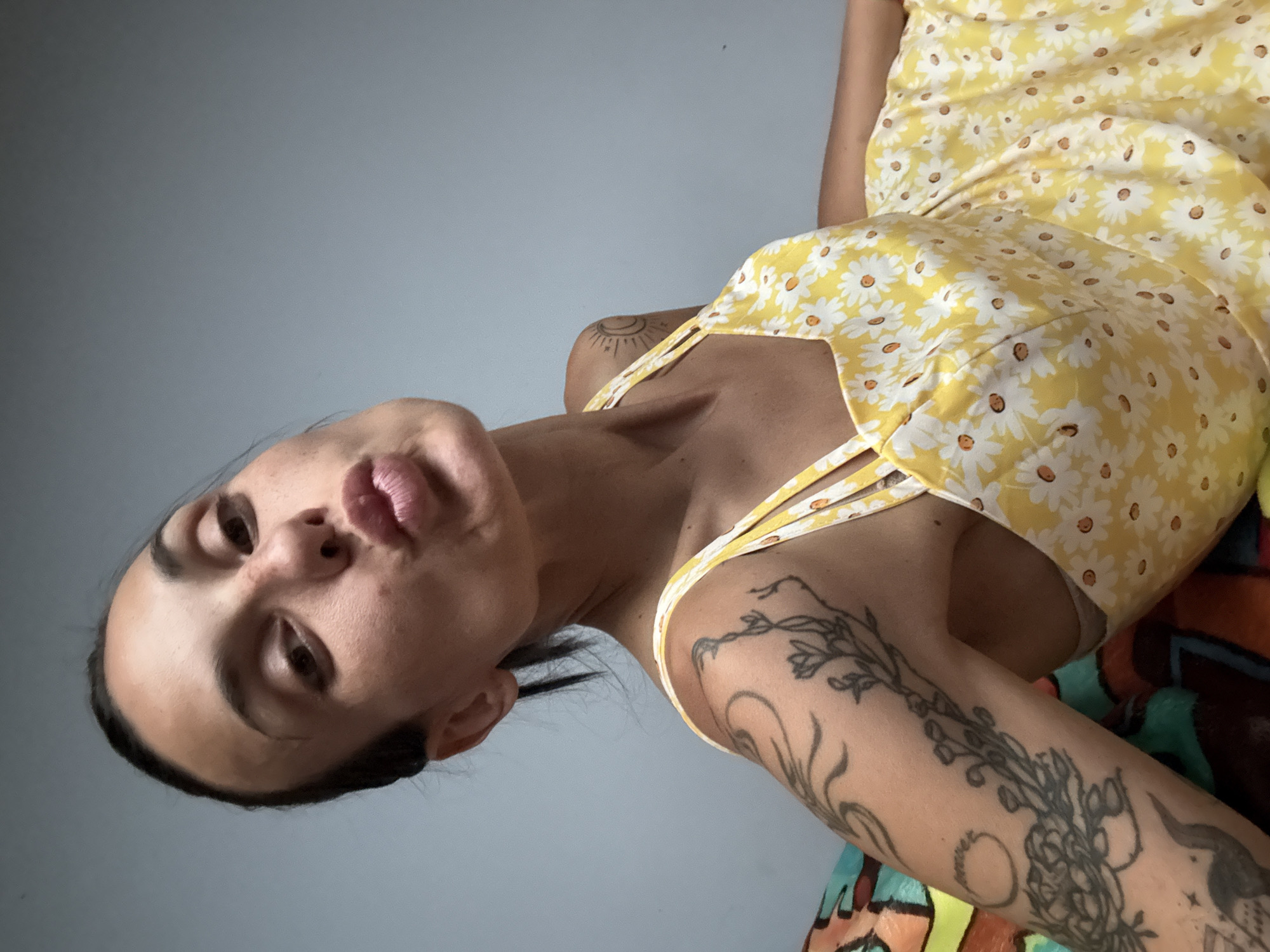A woman with dark hair pulled back is leaning towards the camera, puckering her lips in a kiss gesture. She is wearing a yellow dress with a daisy print and has tattoos on her arms. The background is a plain, light blue wall. The image is a close-up, upper body shot.