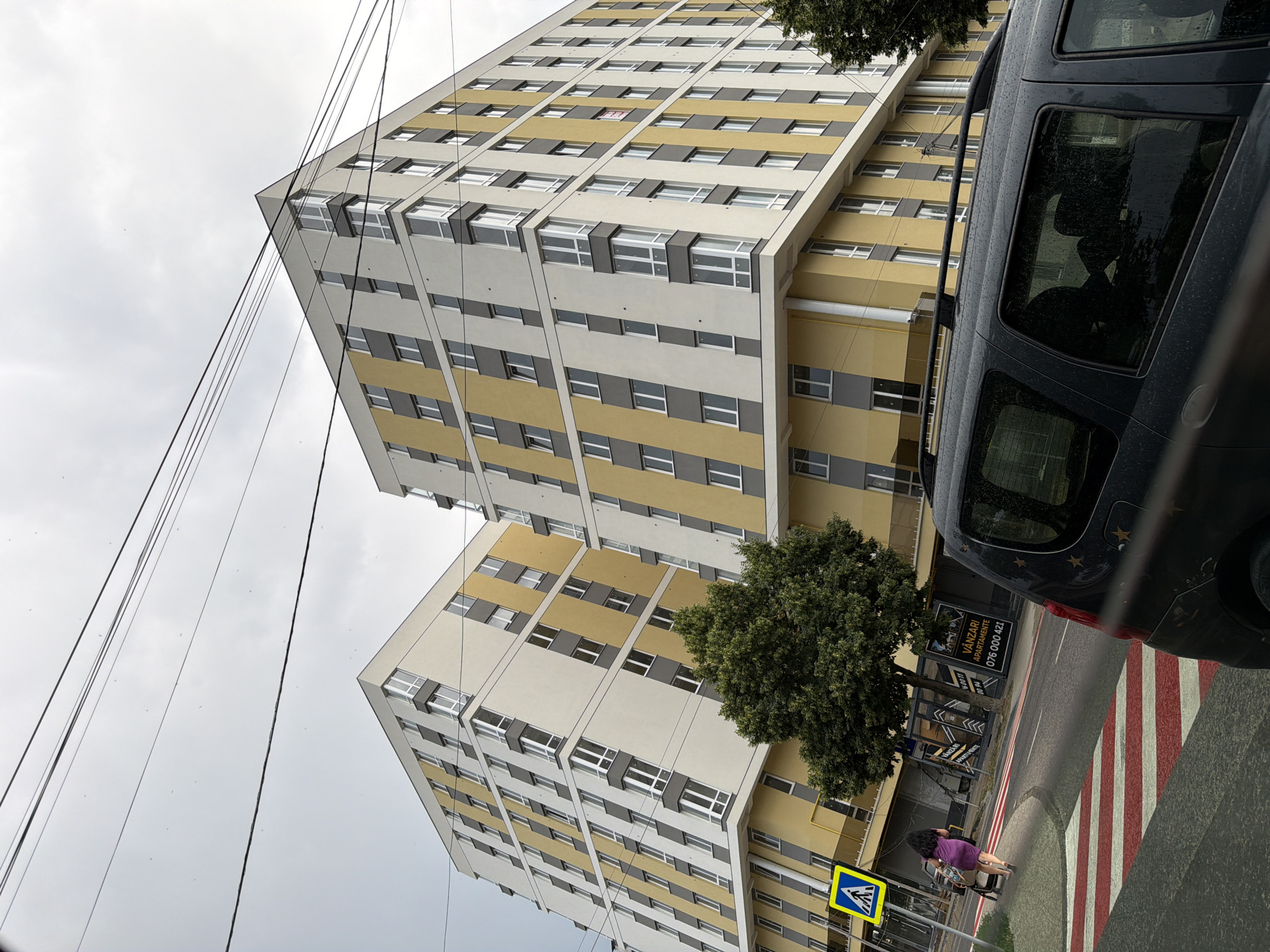 In Strășeni, Moldova, two modern apartment buildings with yellow and white facades stand under an overcast sky. Electrical wires crisscross the sky above the structures. In the foreground, a dark-colored SUV is parked on a street marked with a pedestrian crossing featuring red and white stripes. A woman in a purple dress pushes a stroller on the sidewalk to the left of the street. A yellow pedestrian crossing sign is visible near the woman. One of the apartment buildings has advertisements for "VÂNZĂRI APARTAMENTE" (Apartment Sales) posted on its lower facade. A mature tree with green foliage is situated between the street and the buildings.
