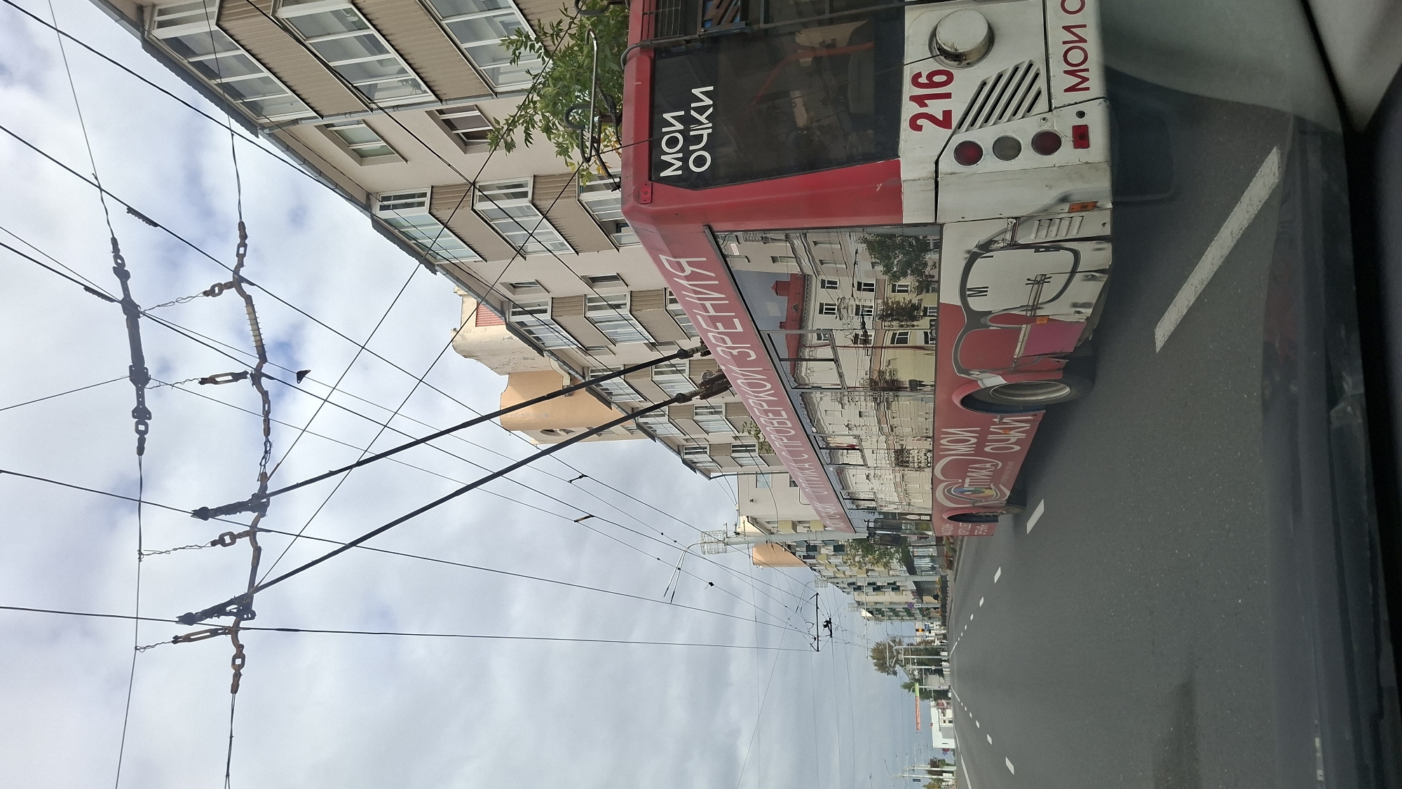 This is a vertical, eye-level, medium shot of a red and white trolleybus driving on a street. The trolleybus has the words "МОИ ОЧКИ" (My Glasses) printed in white letters on its back and side. The number "216" is also visible on the back of the trolleybus. Buildings with multiple windows and balconies line the street. Overhead, a complex network of wires for the trolleybus is visible against a cloudy sky. The scene appears to be a city street in Vitsebsk, Belarus, based on the Cyrillic text on the trolleybus and the architectural style of the buildings. The image is captured from inside a vehicle, as indicated by the reflection in the lower part of the frame.