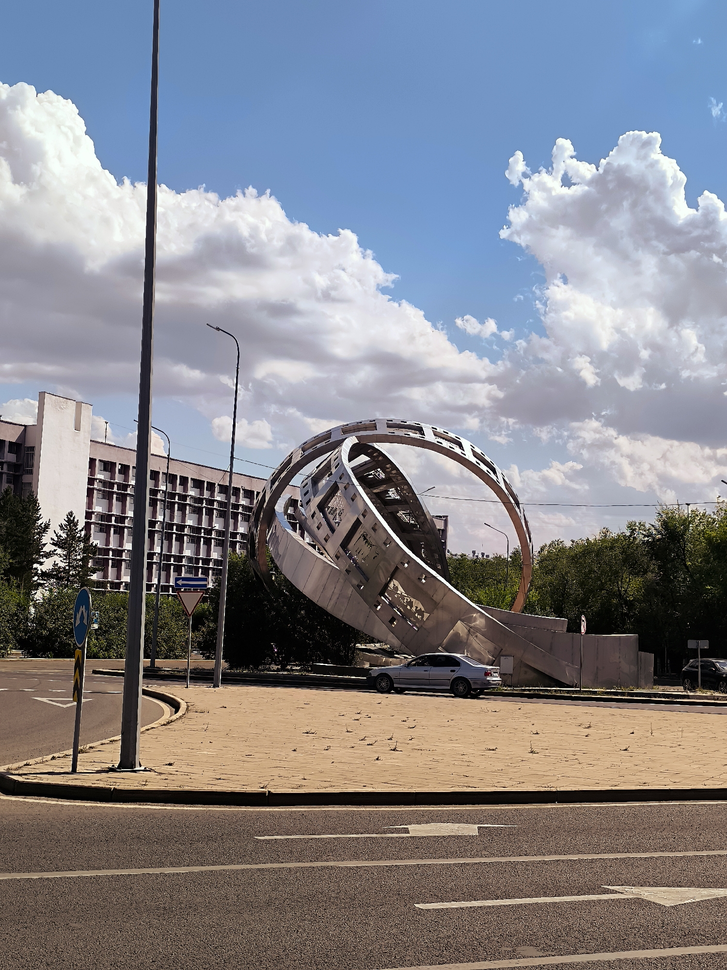In Qarağandı city administration, Kazakhstan, a striking contemporary sculpture dominates the foreground of the image. The sculpture, crafted from polished metal, features interlocking rings and geometric shapes, appearing to twist and curve in a dynamic, abstract form. It is situated in a paved circular area, likely a roundabout or plaza, with a light-colored, textured surface.
To the left, a tall, dark lamppost stands prominently, casting a subtle shadow. Beside it, a light blue circular traffic sign with a white arrow indicating a left turn is visible. Further left, the facade of a multi-story building is seen, characterized by its pale exterior and a repeating pattern of windows with dark frames, bordered by a distinct pinkish-purple trim.
Behind the sculpture and building, a dense line of green trees forms a natural backdrop. The sky above is a vibrant blue, punctuated by large, fluffy white cumulus clouds, suggesting a clear, fair-weather day. In the middle ground, a silver sedan is parked on the paved area near the sculpture. The foreground shows a section of asphalt road with white directional arrows painted on it. The scene appears to be an urban public space, showcasing a blend of modern architecture, public art, and natural elements.
