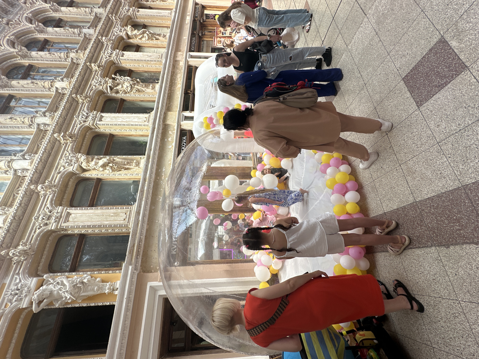 The image captures a bustling scene within the ornate Odesa Passage in Odesa, Ukraine, a multi-story classical revival architectural structure. In the foreground, a blonde woman in a vibrant red sleeveless dress stands with her back mostly to the camera, observing a large, clear inflatable dome. To her right, a young girl with dark braids, wearing a white sleeveless top and shorts, also looks towards the dome.
The central feature of the midground is a transparent inflatable dome, serving as a play area, filled with numerous pink, white, and yellow balloons. Inside, several children are visible, actively playing amongst the balloons; one girl in a patterned dress is prominent. To the immediate right of this dome, a smaller, white inflatable structure is present, also adorned with balloons.
Further to the right, a woman in a beige two-piece outfit stands facing the dome, carrying a light-colored shoulder bag. Next to her, a group of three women are engaged in conversation: one wears a blue jumpsuit, another a black tank top with ripped light-wash jeans, and the third a white long-sleeve top with light-wash jeans.
The background reveals the grandeur of the Passage's interior, characterized by highly decorative building facades. These multi-tiered structures feature numerous windows flanked by elaborate architectural details, including engaged columns, intricate carvings, and statues of draped female figures and possibly atlantes supporting upper balconies. Storefronts are visible along the passage, with signs bearing the name "KVITKOVA." The floor is tiled with light-colored speckled squares, interspersed with a darker, rectangular tiled section in the central walkway.