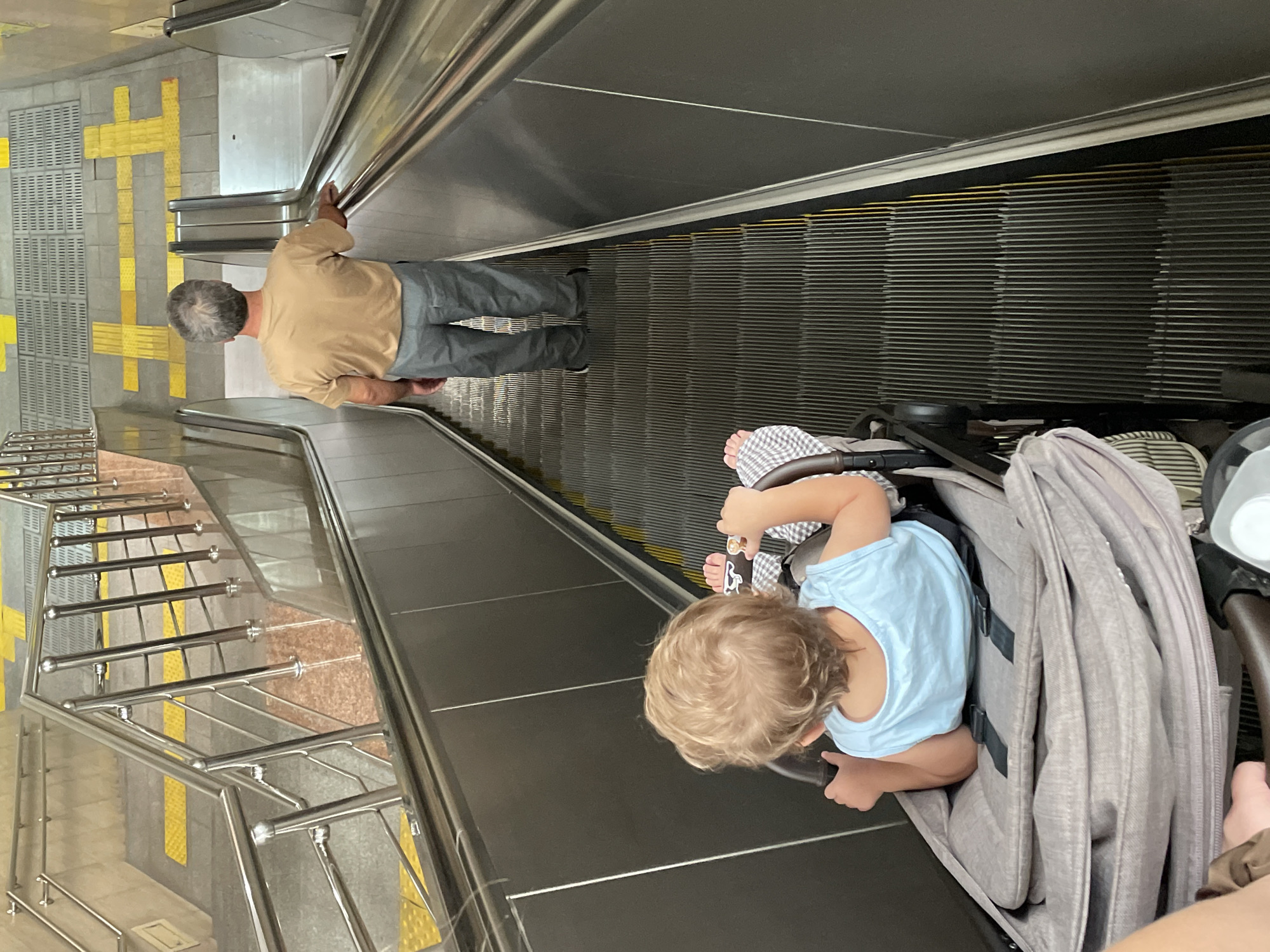 An overhead view captures a downward-moving escalator in Almaty, Kazakhstan. A man with gray hair, wearing a tan t-shirt and gray pants, stands on the escalator, holding onto the handrail. In front of him, a toddler sits in a stroller, facing away from the camera, wearing a light blue tank top and checkered pants. The escalator has metal steps with yellow safety lines, and stainless steel handrails line both sides. The surrounding environment includes textured flooring with yellow tactile paving at the top of the escalator.