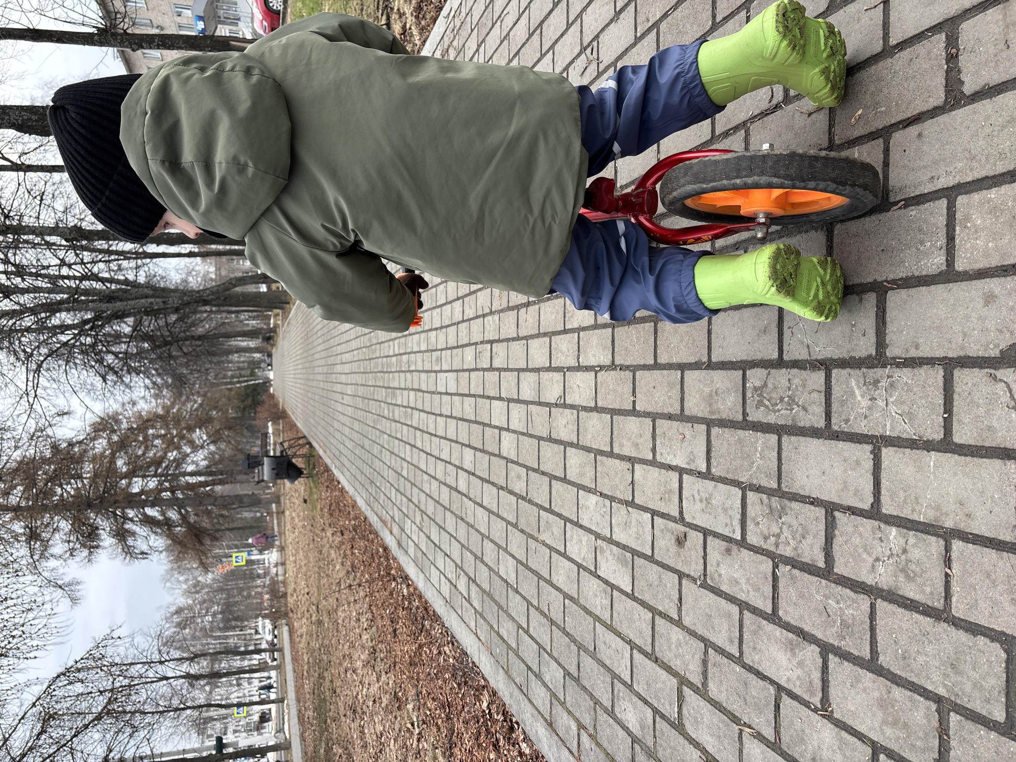 A child with a black beanie, green jacket, blue pants, and green rubber boots is riding a red balance bike with orange wheels on a brick sidewalk. The sidewalk runs alongside a street with trees on the left. The location is Chaikovsky, Russia.