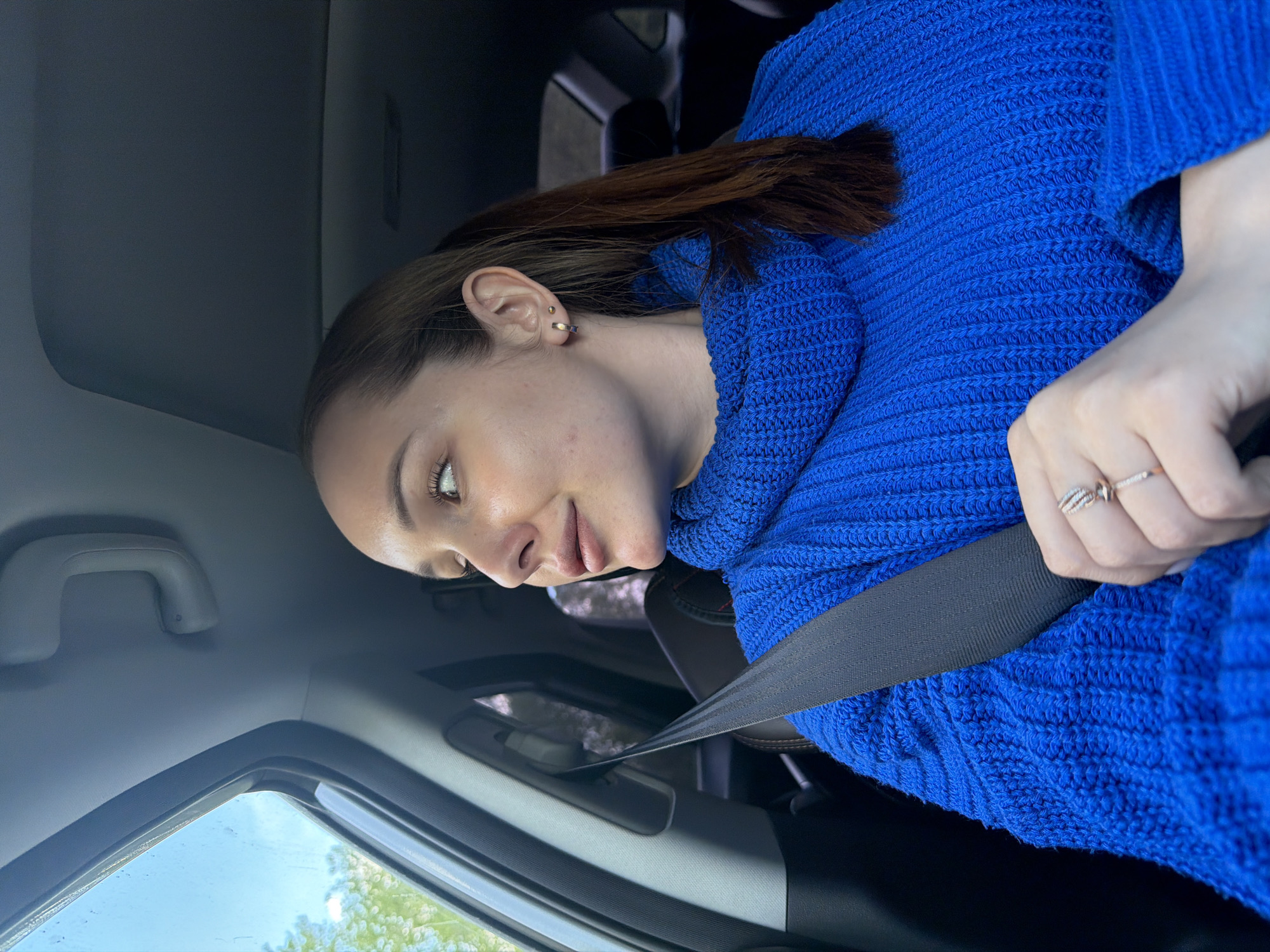 A young woman is seated in the driver's seat of a car, wearing a blue sweater and a seatbelt. She has brown hair pulled back and is looking slightly to her left. She is wearing rings on her left hand. The interior of the car is visible, including the car's window and the dashboard. The location is unknown and there is no indication it is in Chaikovsky, Russia.