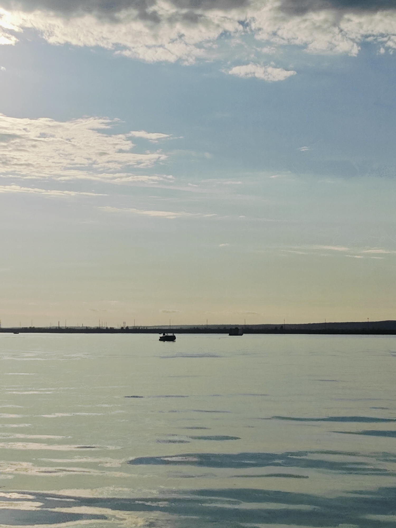 The image depicts a serene body of water, likely a river or reservoir, with a distant, silhouetted shoreline. The sky is a soft gradient of blues and whites, with scattered clouds that catch the light. A small boat with a dark silhouette is visible on the water in the middle distance. The shoreline is lined with what appear to be industrial structures and transmission towers, suggesting an urban or industrial area. The water's surface is calm, with subtle ripples reflecting the sky. The overall impression is one of tranquility and a vast, open landscape. This scene is consistent with views of the Kama River in Chaikovsky, Russia, which is known for its hydroelectric power station and surrounding industrial infrastructure.