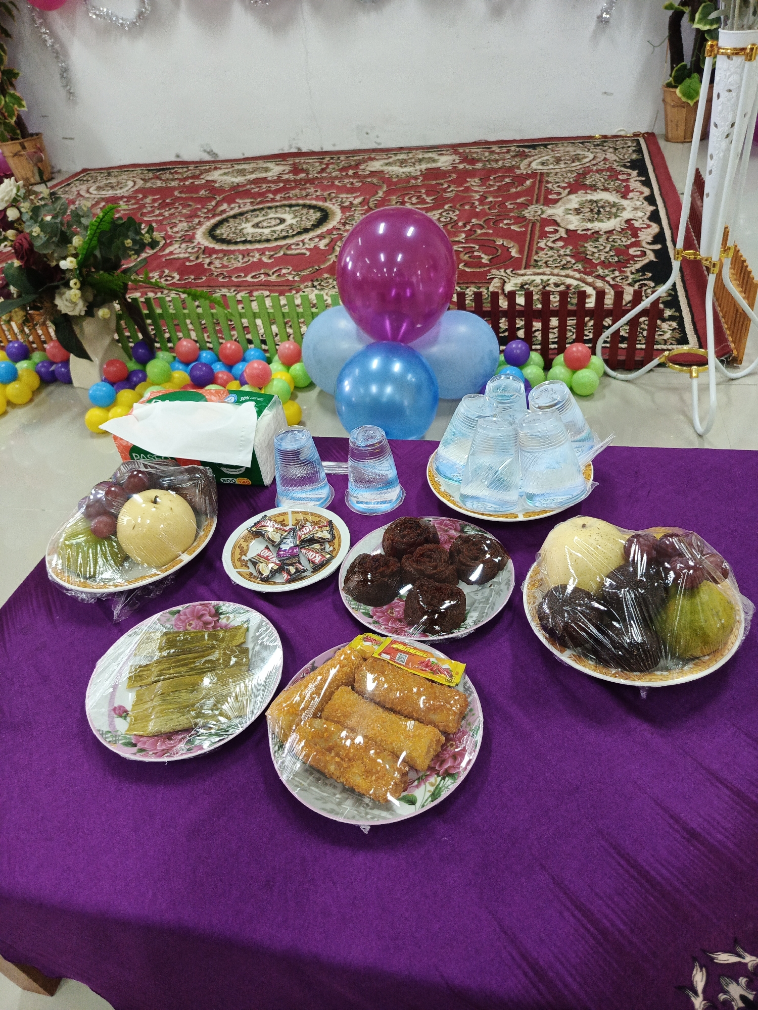 The image depicts a table laden with food items and decorations, suggesting a celebration or gathering. The table is covered with a purple tablecloth, and on it are several plates of food, including fruit, pastries, and what appear to be fried snacks. There are also stacks of clear plastic cups and a box of tissues. In the background, colorful balloons and a patterned rug are visible, along with a floral arrangement in a vase. A low, green fence separates the table area from a collection of small, multi-colored balls, possibly from a ball pit. The overall impression is one of a festive event, likely a birthday party or similar occasion, held indoors. The presence of the rug and the general decor suggests a domestic or community hall setting. Given the mention of Lhokseumawe, Indonesia, the scene is likely within a residence or a local venue in that city. No people are visible in the image.
