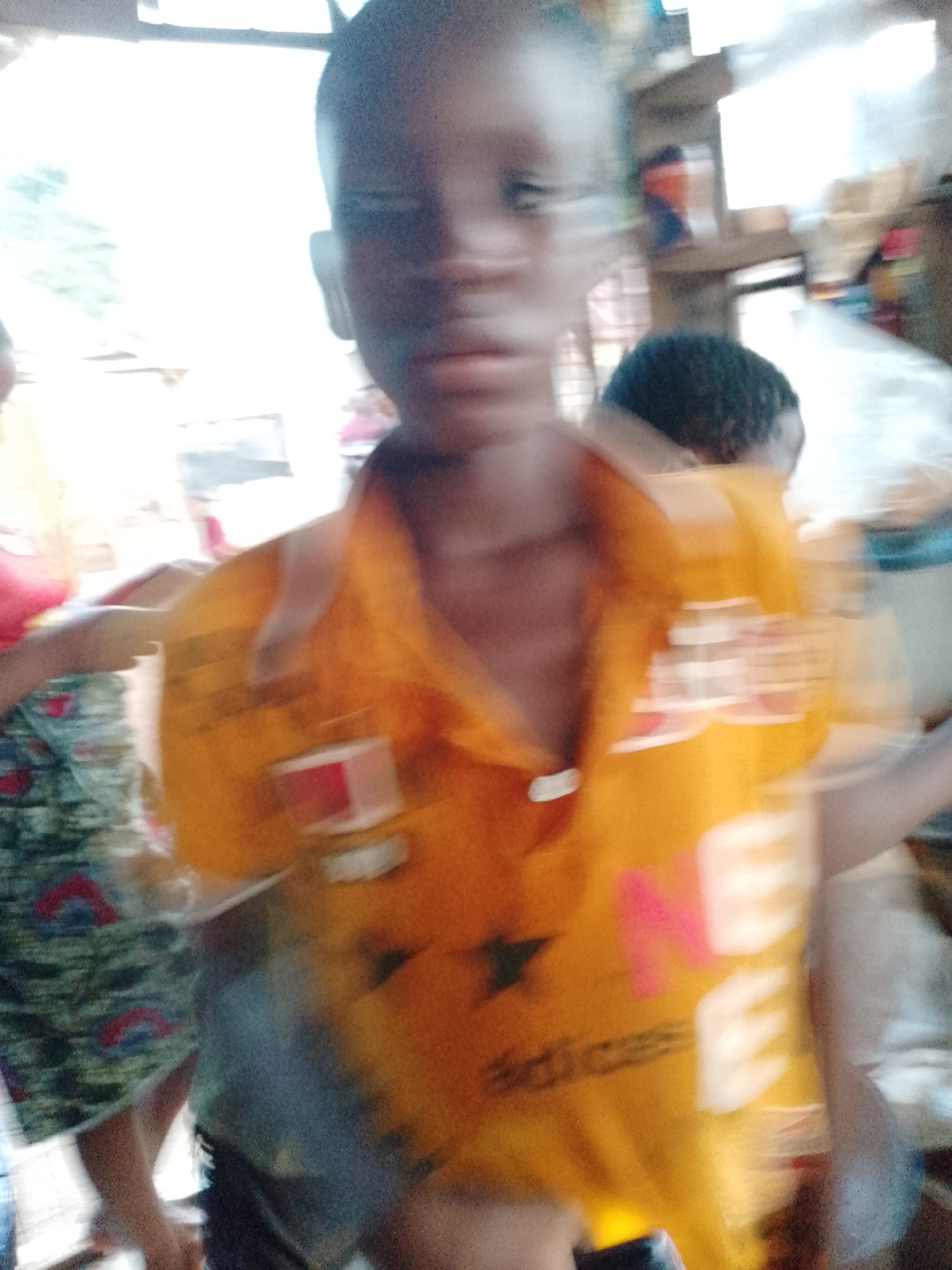 A blurred image depicts a child in Akwanga, Nigeria, wearing an orange t-shirt with pink and white lettering. Another person stands to the left, wearing a patterned skirt and red top. Another person with short, dark hair is visible behind the child. The background suggests an indoor setting, possibly a market or shop, with various items visible.