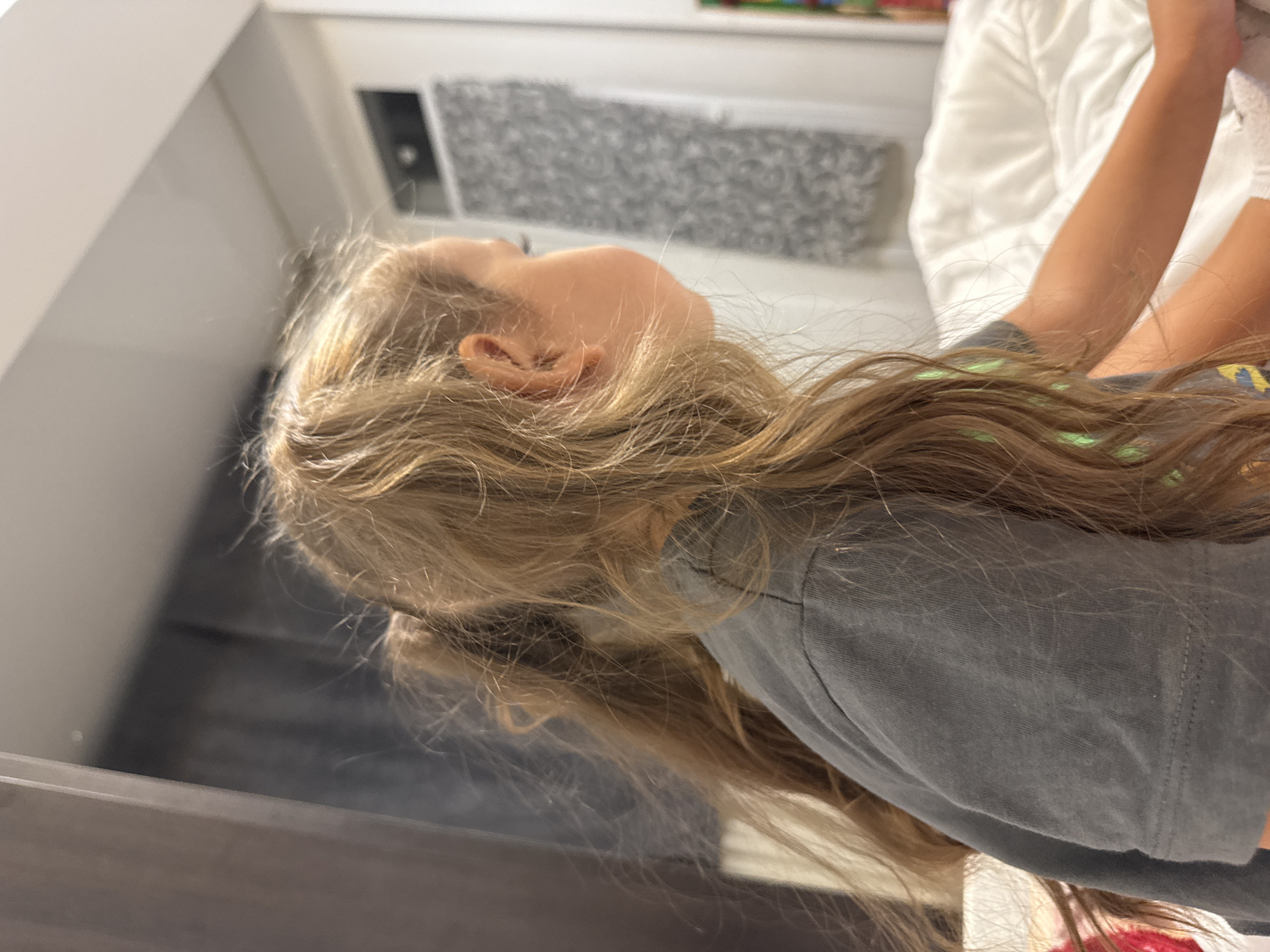 The image shows a close-up profile view of a young girl with long, wavy blonde hair tied in two pigtails. She is wearing a dark grey t-shirt. Her head is turned to the right, and she is looking off-camera. The background appears to be a bedroom with a white duvet or bedspread and a dark curtain on the left. A patterned wall covering or artwork is visible in the background on the right. The lighting suggests an indoor setting, possibly during the day or with artificial light. There is no information in the image to suggest a location in Chișinău, Moldova.