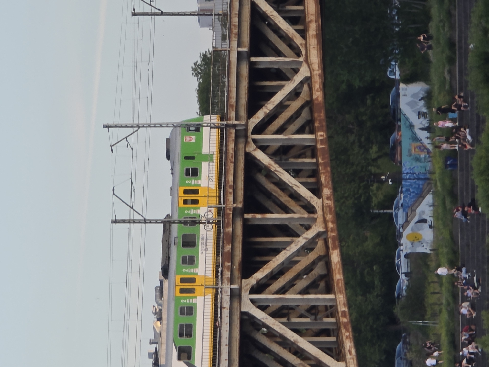 The image shows a green and yellow train on a bridge in Warsaw, Poland, likely crossing the Vistula River. The train has the number "2" visible on its side, as well as what appears to be a Polish insignia. The bridge itself is a steel truss structure, showing signs of rust. Below the bridge, there is a grassy embankment where people are sitting on steps, some in groups. Further in the background, cars are parked along a road, and there is a mural on a wall. The sky is clear and a pale blue.